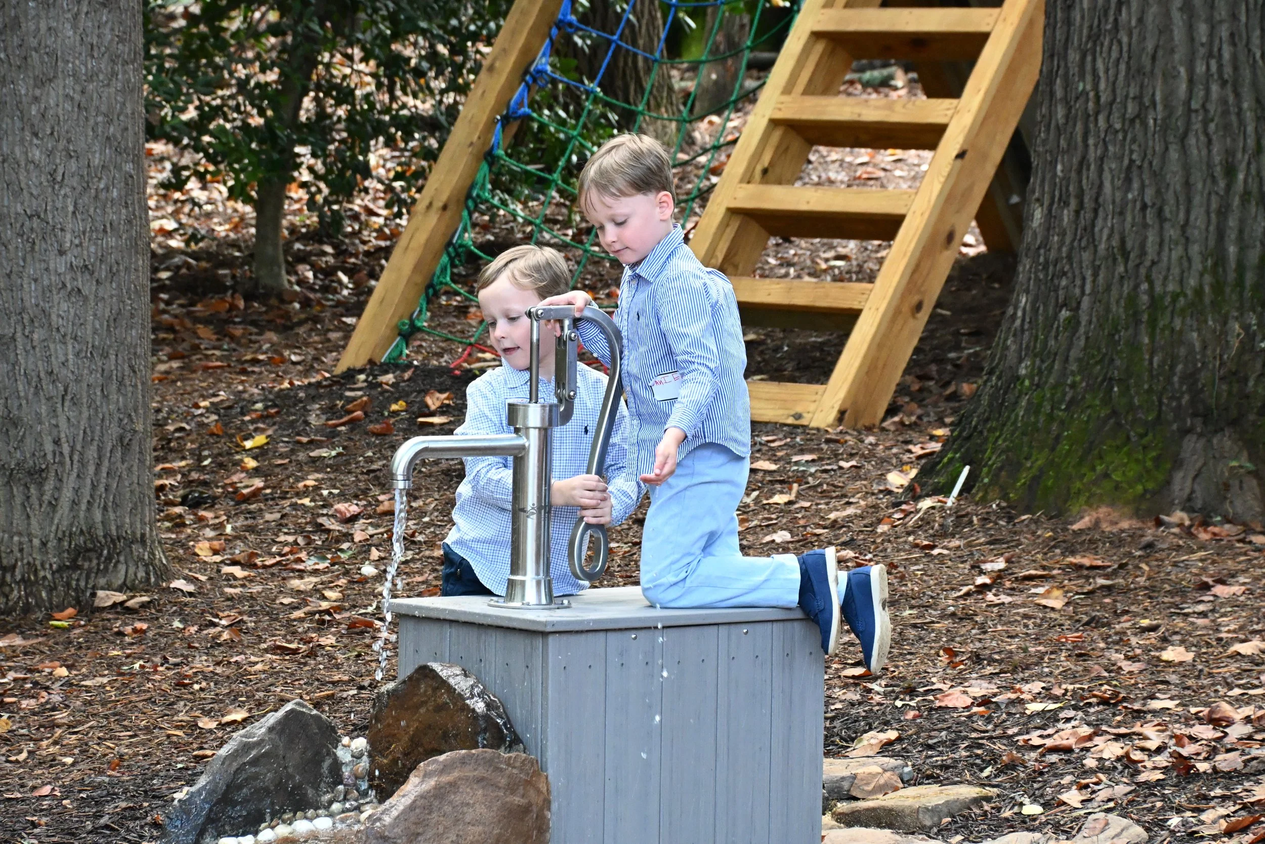 Two young boys in blue plaid shirts playing at a water fountain outdoors in a wooded park, with a wooden staircase and climbing rope in the background.