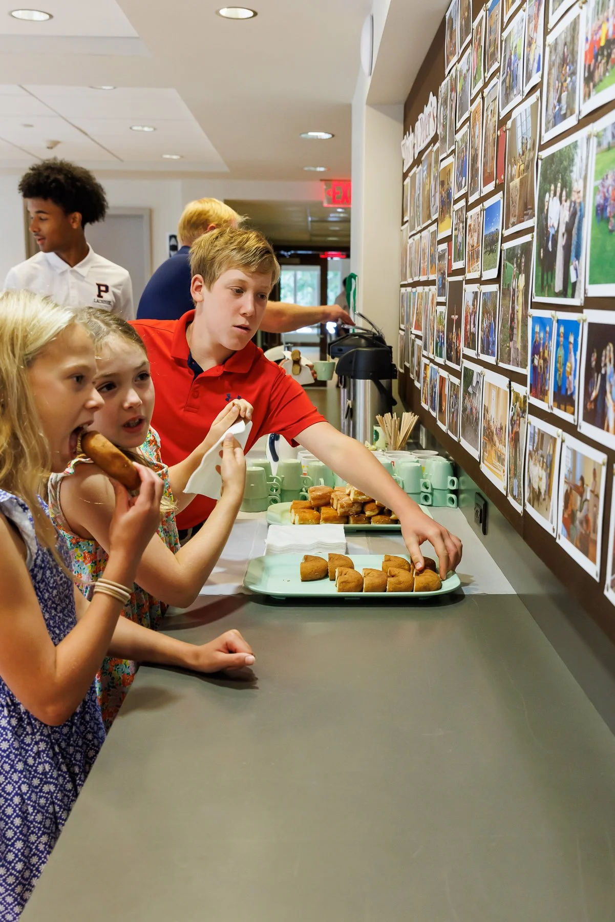 Children at a buffet table, with a girl eating a sausage roll and a boy reaching for a pastry, while others look at a wall of photos.