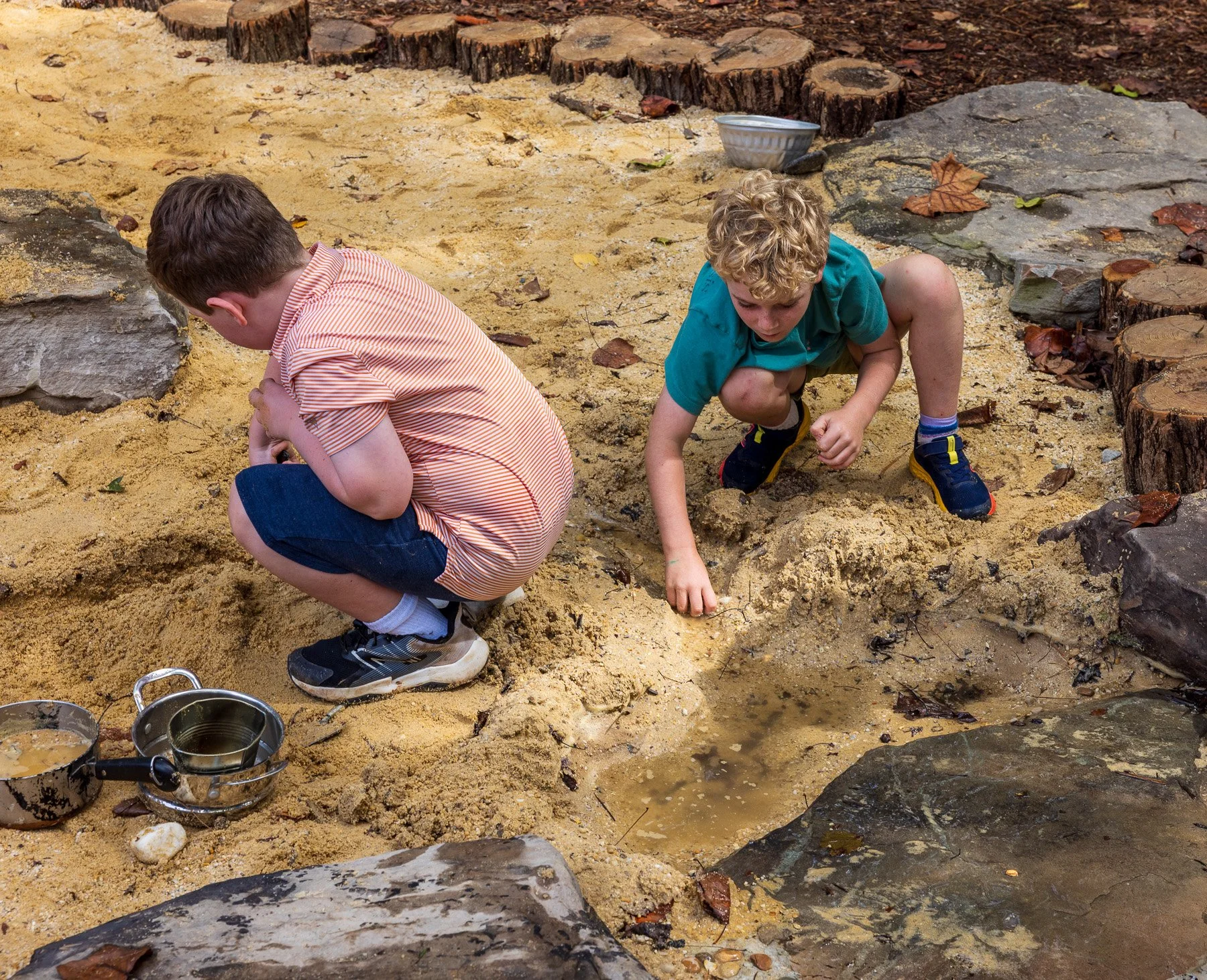 Two young boys playing in a sandy area surrounded by large rocks and tree stumps, with toys and animal footprints in the sand.