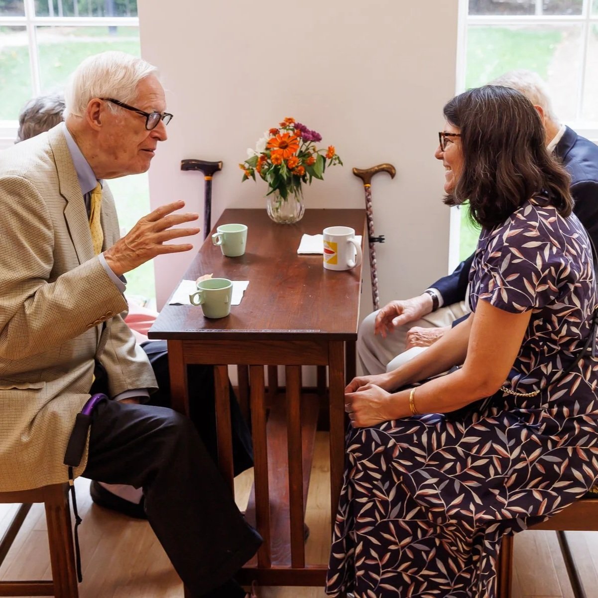 Older man and woman having a conversation at a wooden table with cups, mugs, and a vase of flowers, in a brightly lit room with large windows.