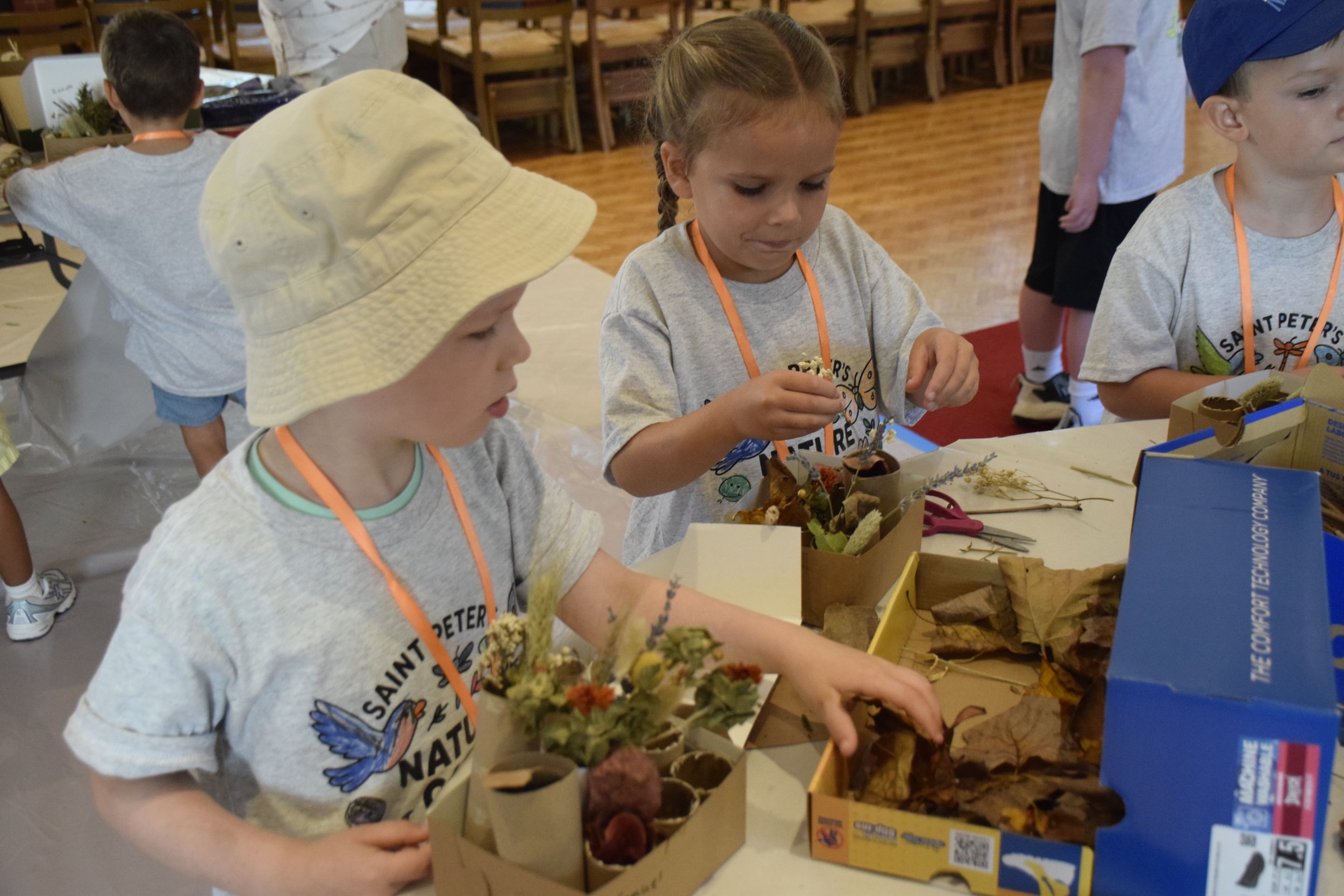 Children participating in a craft activity at a table, creating arrangements with dried flowers and leaves. They are wearing light gray T-shirts and orange lanyards, in a room with wooden chairs and tables.