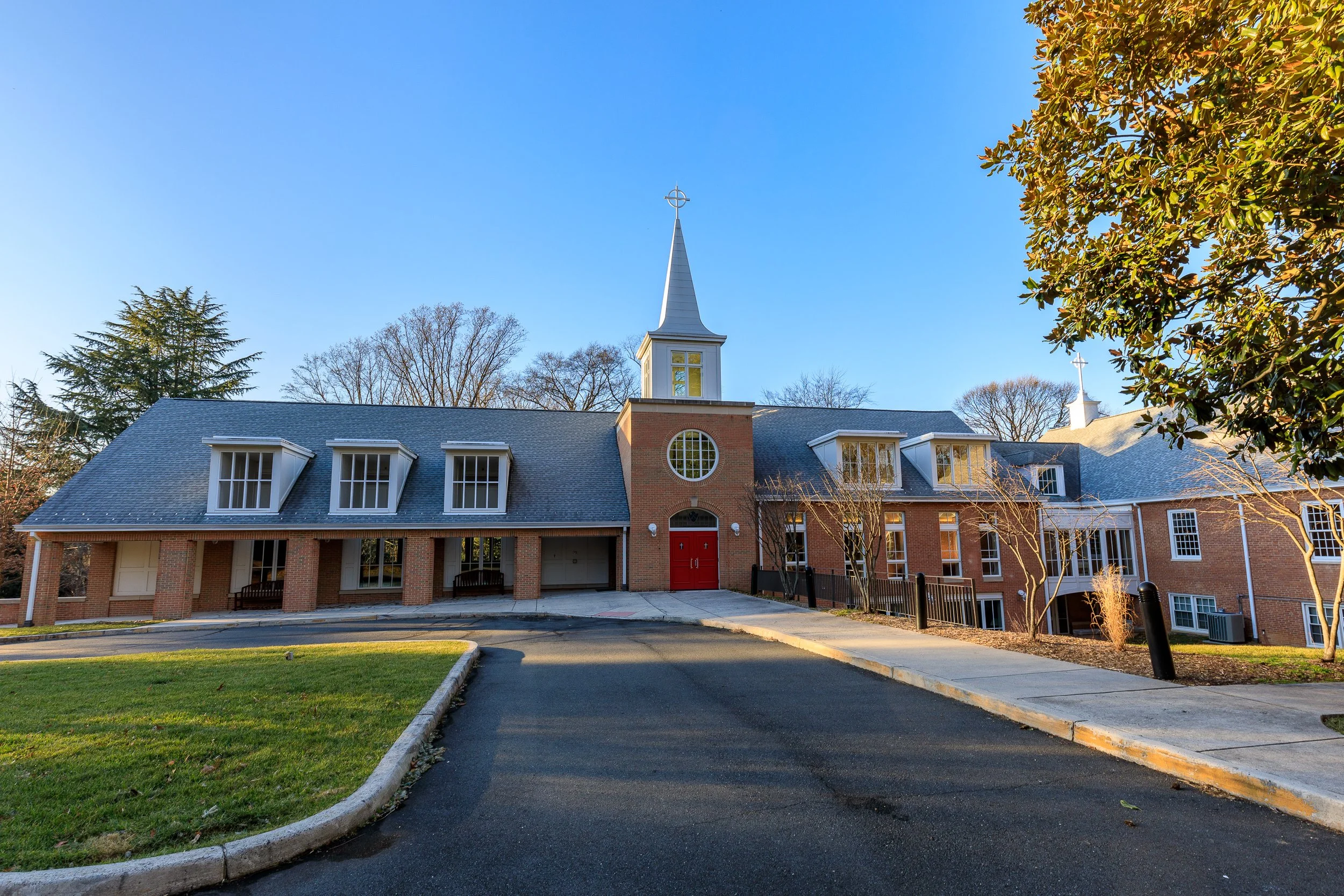 A church building with a tall steeple and crosses on top, red doors, and several windows, surrounded by trees and a paved driveway under a clear blue sky.