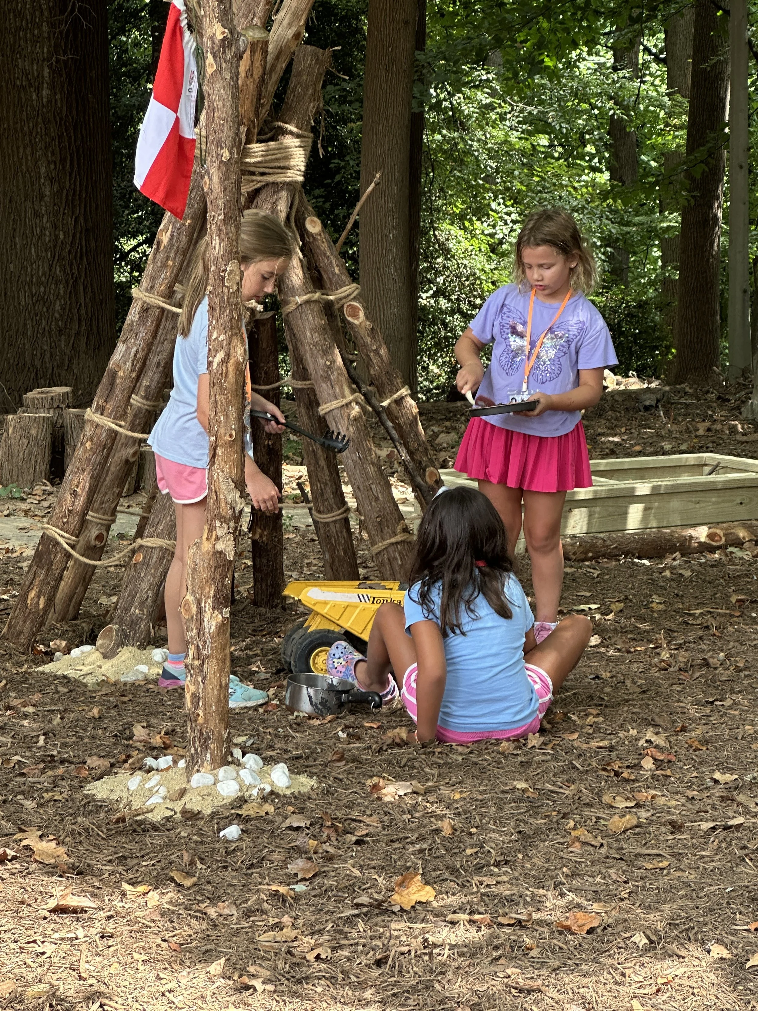 Three young girls playing outdoors in a wooded area, near a makeshift teepee made of wooden sticks. Two girls are standing, one holding a tablet and the other a small gardening rake, while the third girl is sitting on the ground with a toy dump truck, with a metal bowl nearby. The setting appears to be a play area with a wooden sandbox in the background.