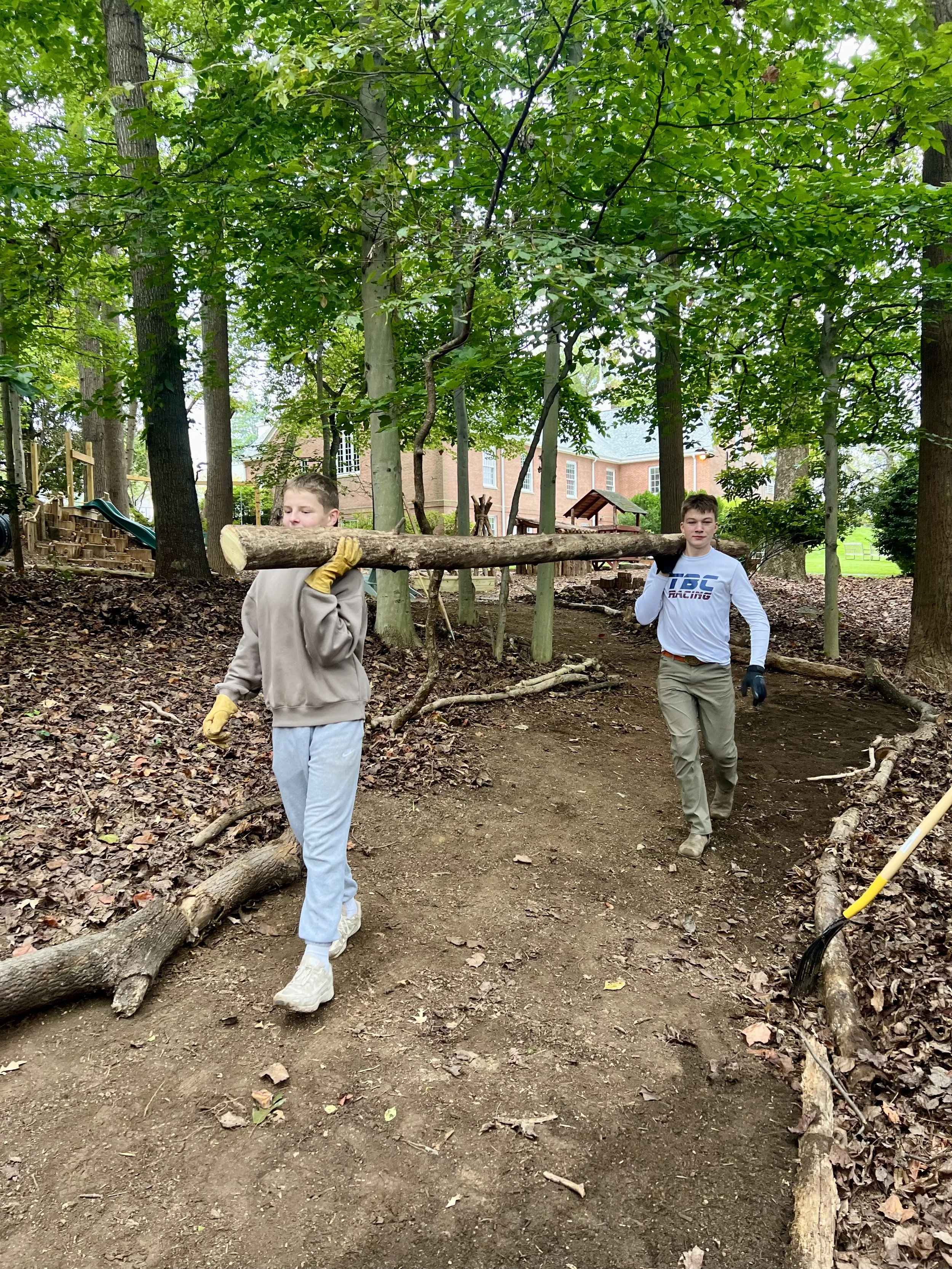 Two young boys work together to carry a large fallen tree branch through a wooded area with a cleared dirt path, with trees and a house in the background.