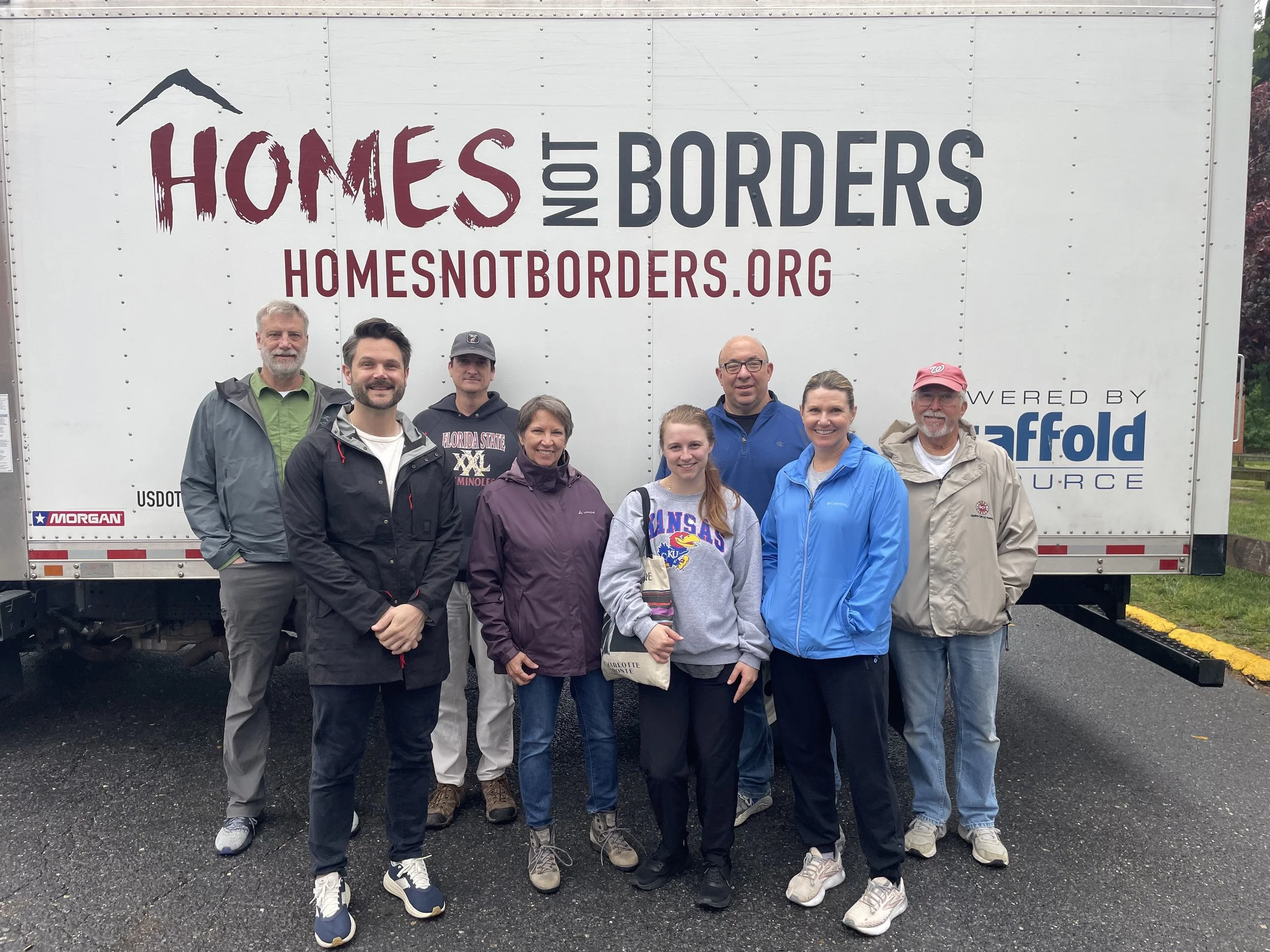 Group of eight people standing outdoors in front of a large truck with the words 'Homes Not Borders' and a website URL. The group appears to be diverse in age and clothing suited for mild weather.