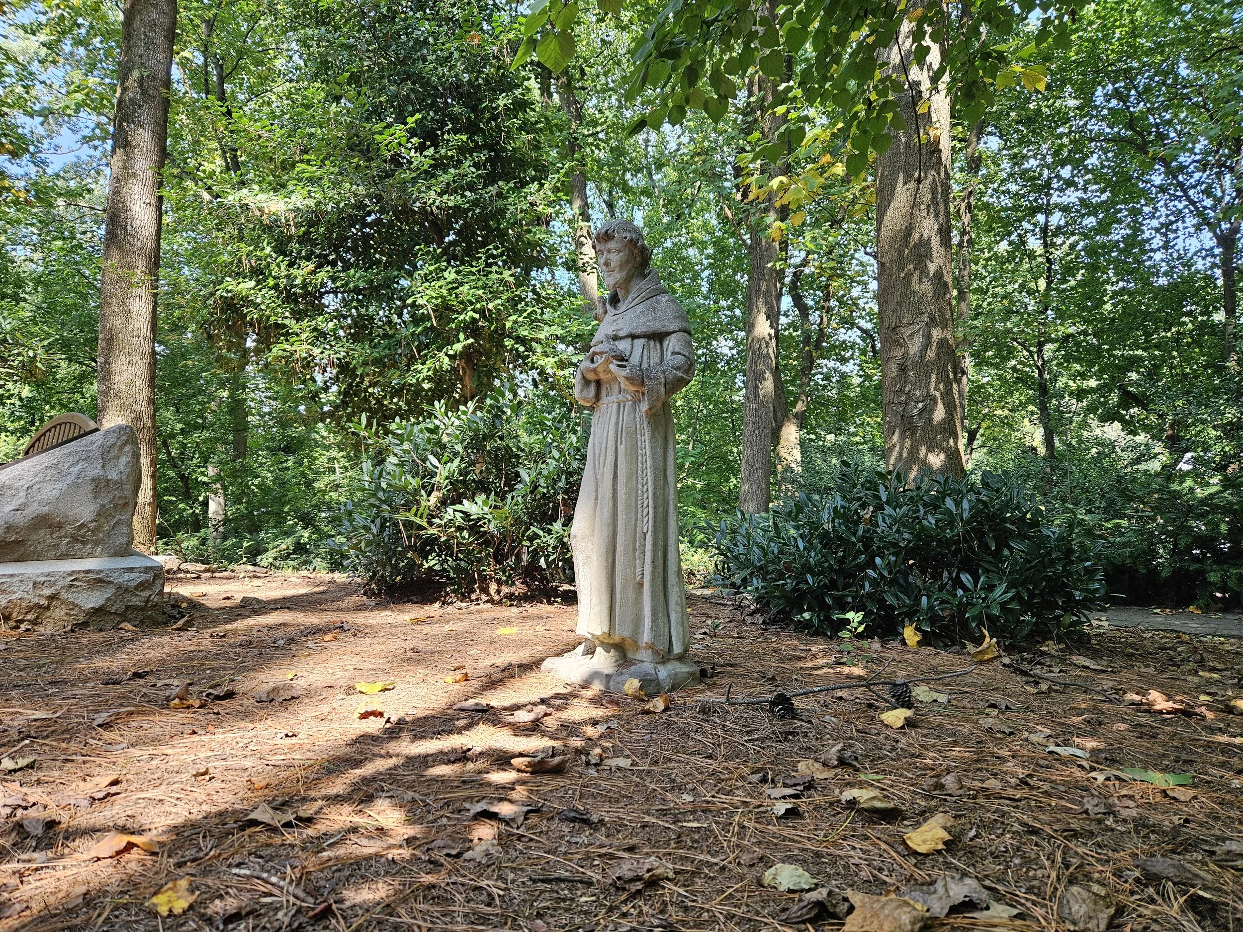A stone statue of a woman in a praying position stands on a forest floor covered with dry leaves, with tall trees and green foliage in the background.