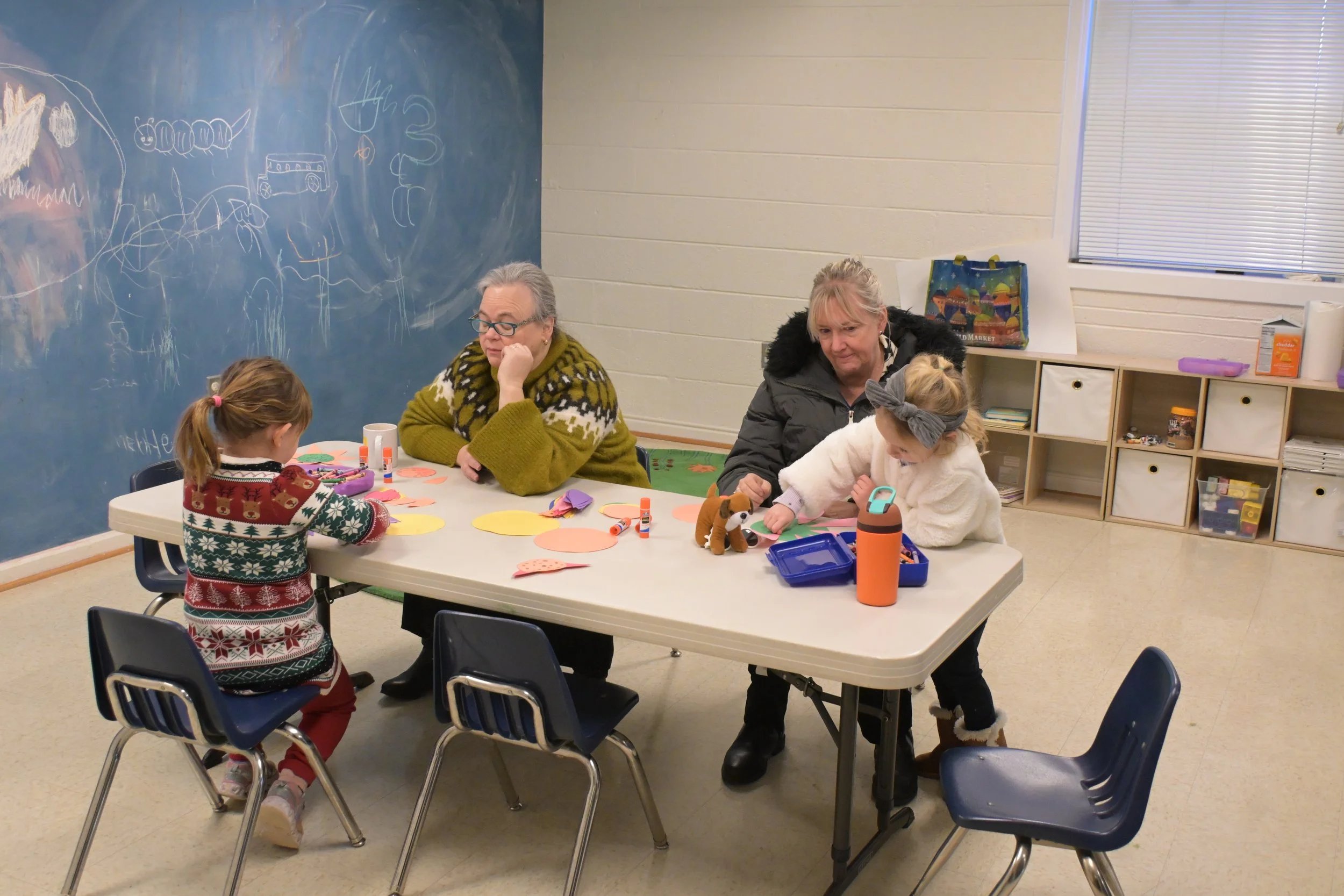 Two adults and two children at a table in a classroom, working on arts and crafts projects with paper, glue, and decorations, with a chalkboard in the background and shelves with supplies.