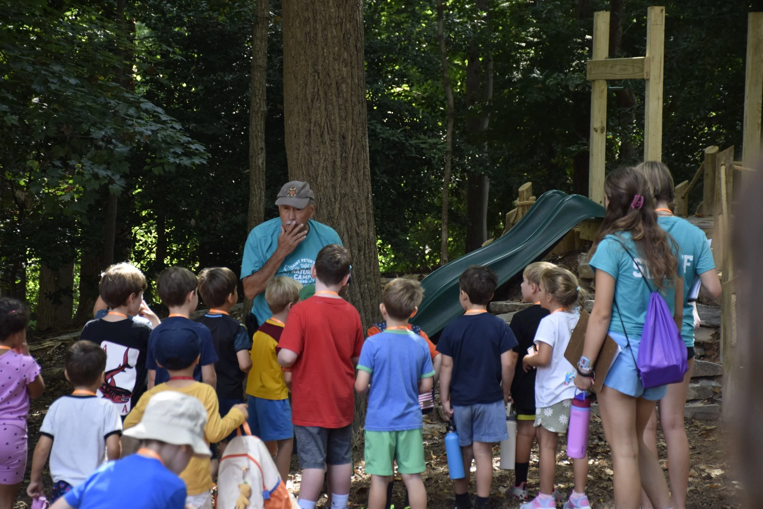 Group of children gathered around a man in an outdoor wooded area, some children have water bottles, and a slide is visible in the background.