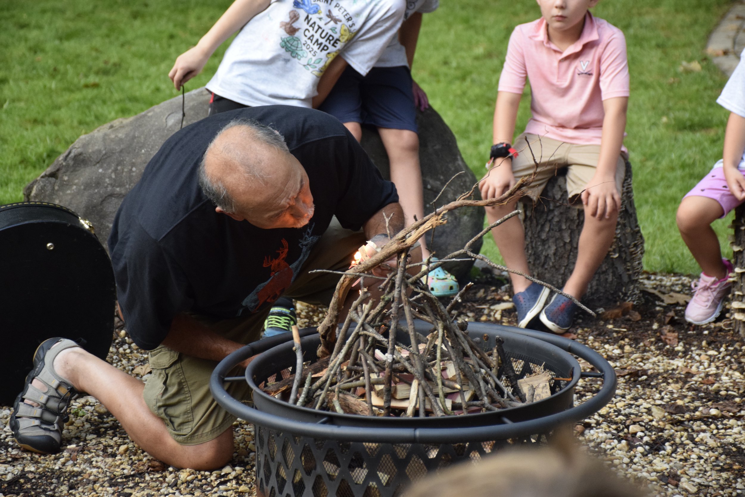 An elderly man crouches down on one knee with a small campfire made of sticks in front of him, surrounded by children sitting on logs and standing nearby outdoors on a grassy area.