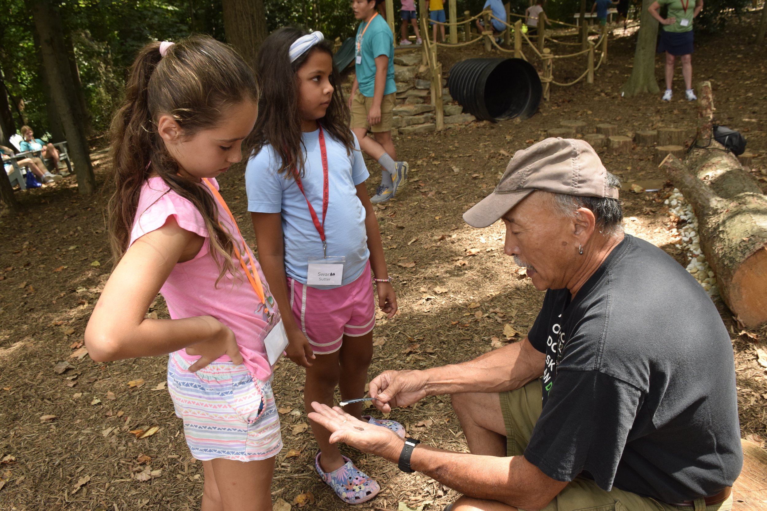 Two young girls with name tags stand in a wooded outdoor area, attentively listening to an older man who is showing them something in his hands. Other children and adults are seen in the background.