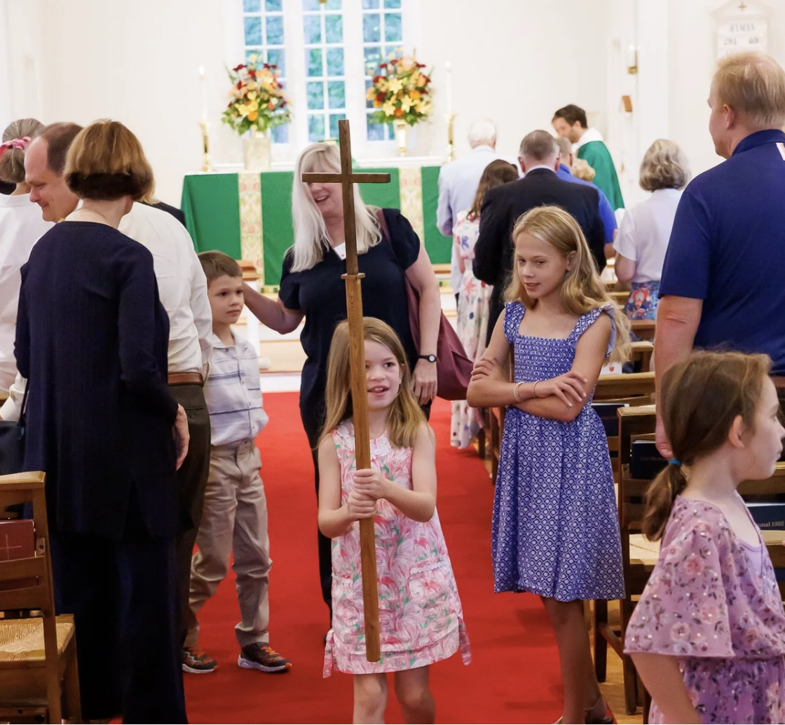 Young girl holding a wooden cross at a church service with several people, including children and adults, gathered inside the church.
