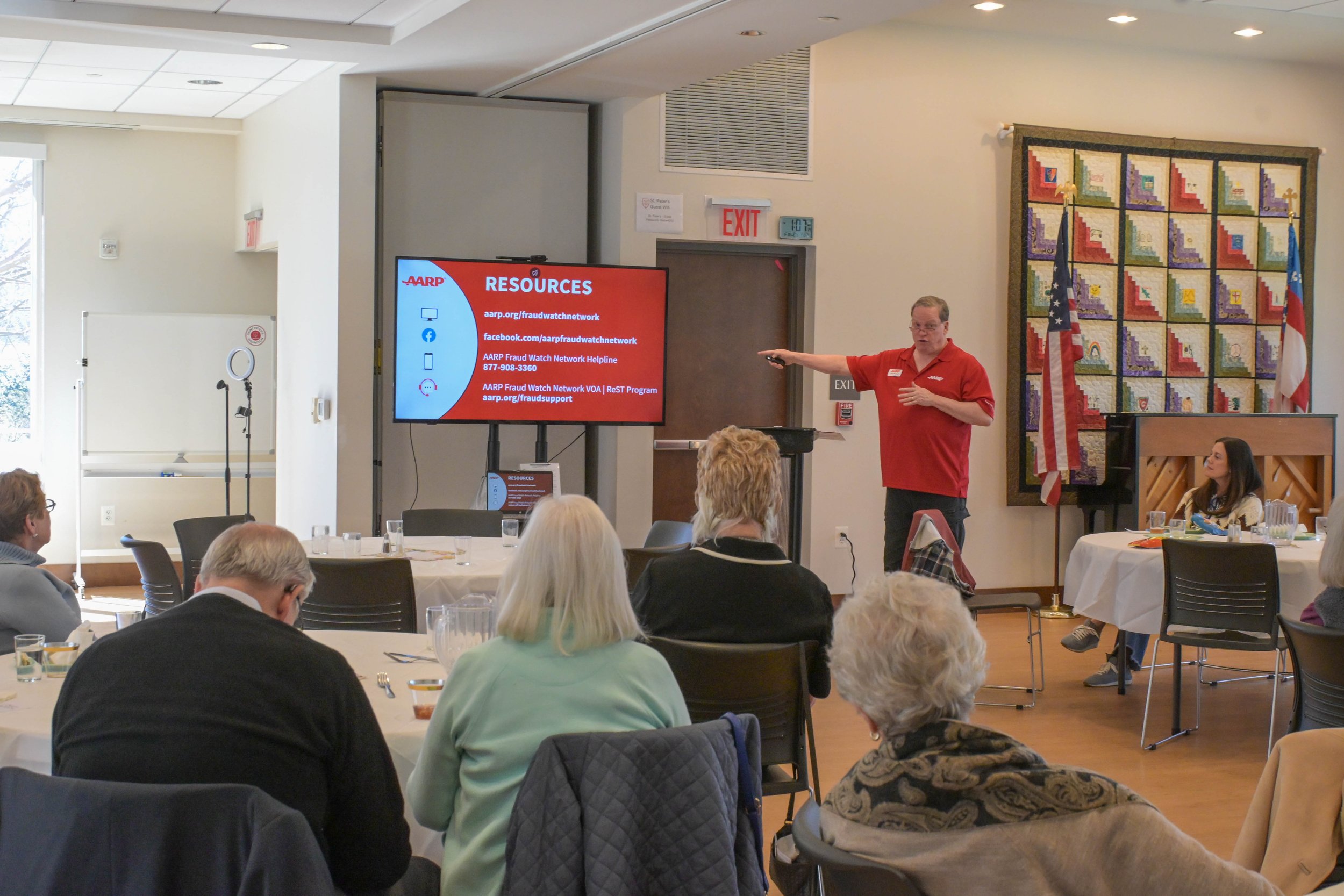 A man in a red shirt giving a presentation to a group of people seated at round tables in a conference room. The man is pointing at a large screen displaying resources related to fraud watch network. The room has flags and a quilt on the wall.