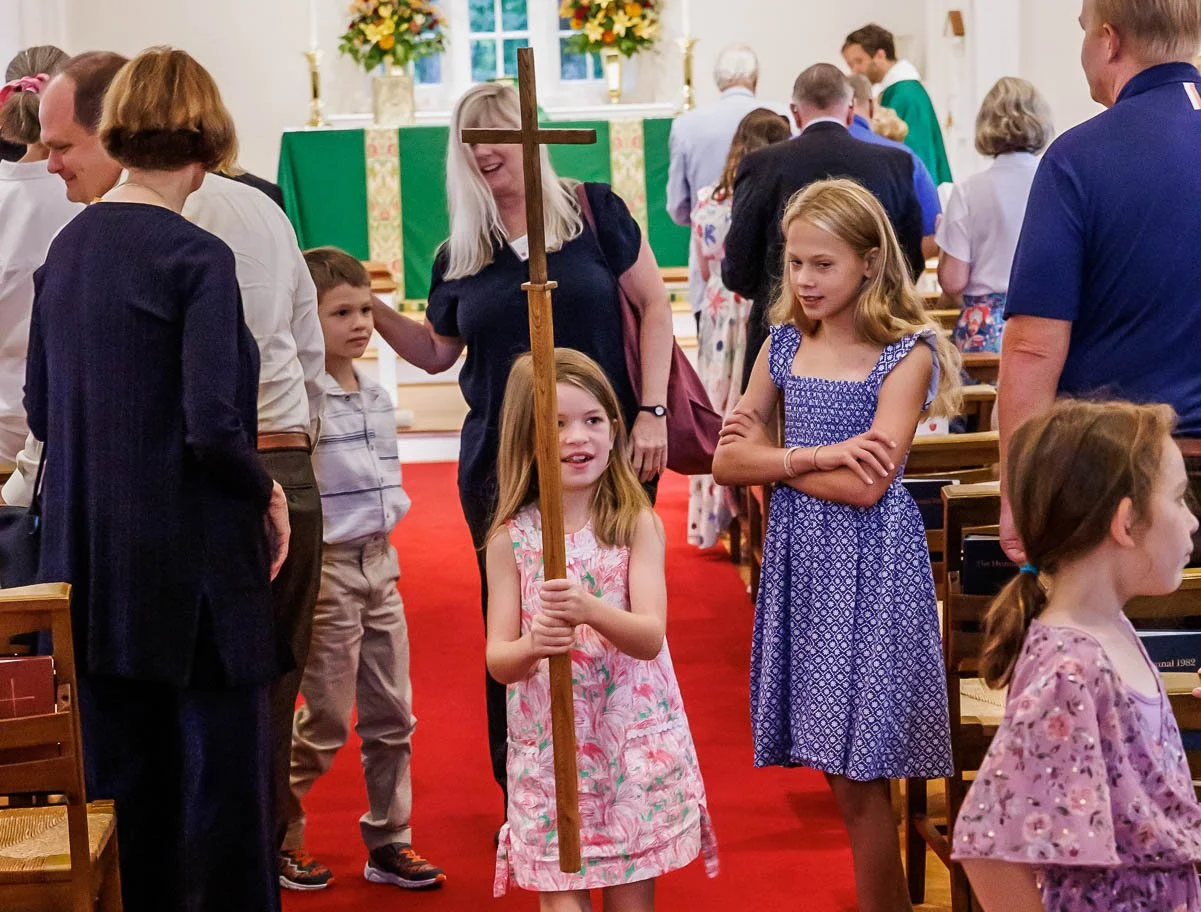 Girl holding a wooden cross in a church during a religious service, surrounded by congregation members.