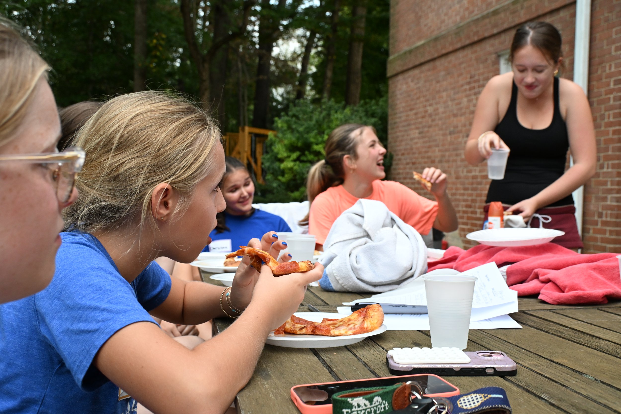 Group of young girls enjoying pizza and drinks outdoors at a wooden table during daytime.