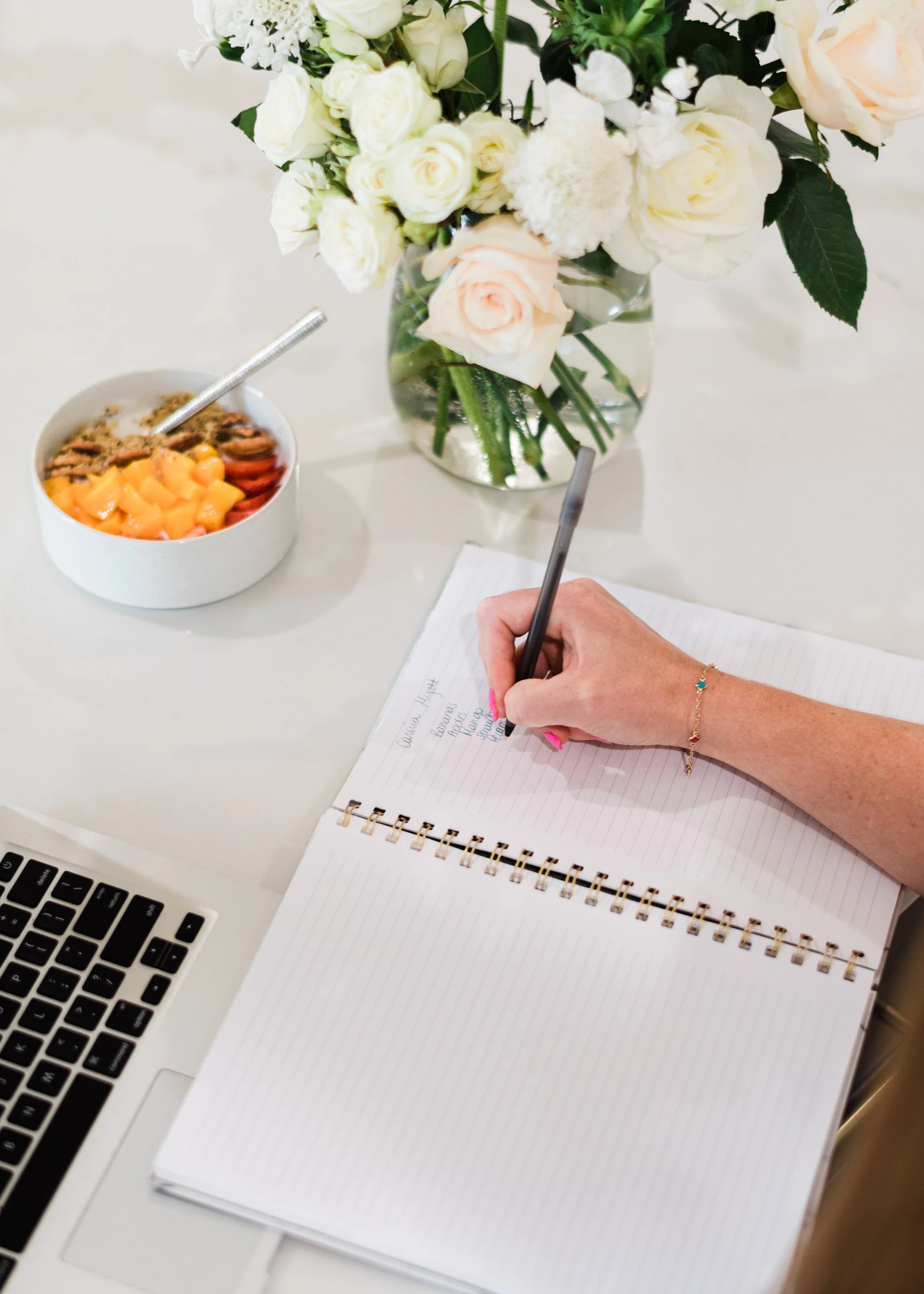 A person writes in a spiral notebook at a desk with a MacBook, a bowl of fruit, and a floral arrangement in a glass vase.