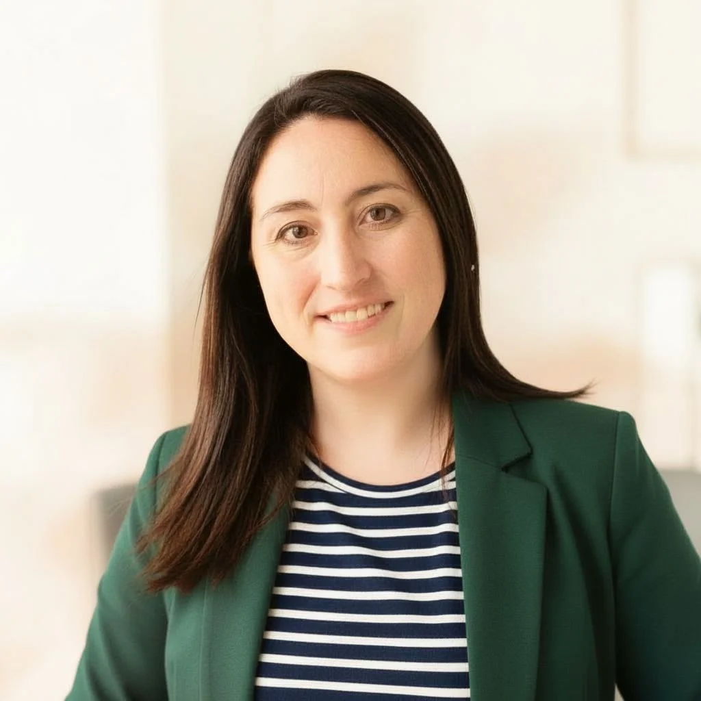A woman with dark brown hair smiling, wearing a green blazer over a striped shirt, standing indoors with a blurred light-colored background.