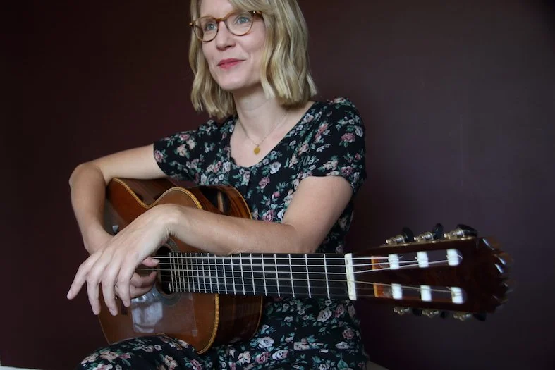 Woman in glasses holding an acoustic guitar against a dark background.