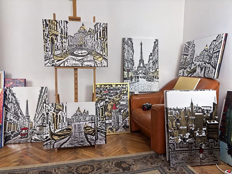 Collection of black-and-white and colored cityscape paintings featuring Paris landmarks, including the Eiffel Tower and Notre-Dame Cathedral, displayed against a white wall with a brown leather couch and a small table nearby.