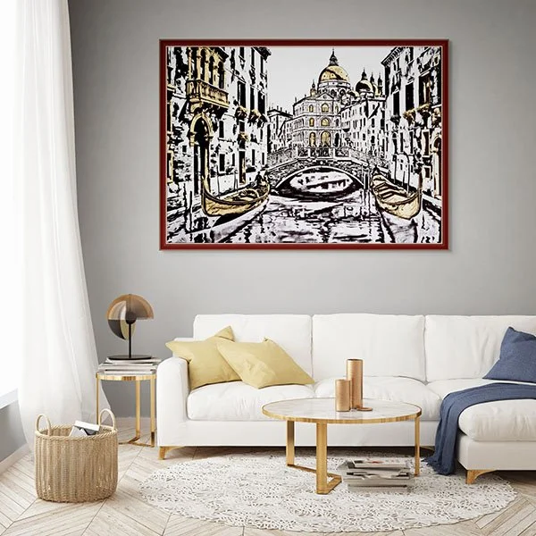 A living room with white sofa, beige and navy pillows, round wooden coffee table, table lamp, and framed painting of a canal scene with buildings and boats, hung on a light gray wall.
