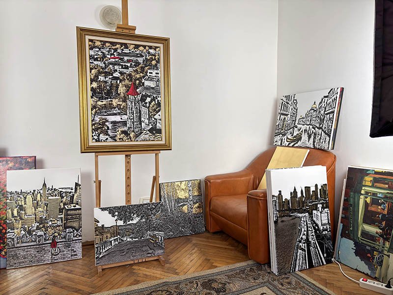 An art studio corner with several cityscape paintings leaning against a white wall and on an easel, a leather sofa, and a hardwood floor.