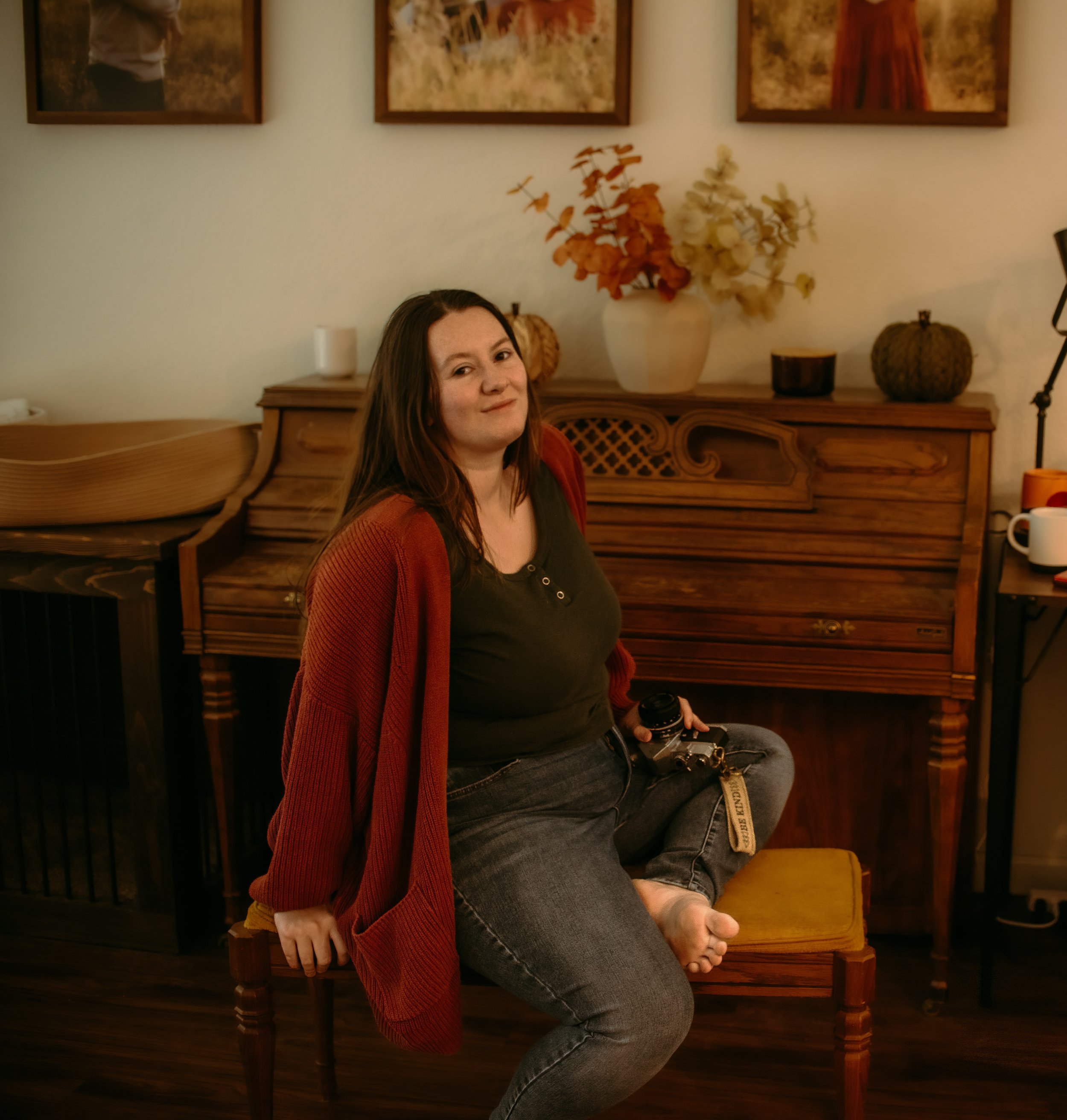 A woman with long brown hair sitting on a wooden bench at a cozy, warmly lit room with autumn-themed decor. She is smiling and wearing a black top and a reddish-brown cardigan. Behind her, there is a piano with dried flower arrangements on top, and framed artwork on the wall. To her right, a person is partially visible, holding a vintage camera and wearing gray jeans.