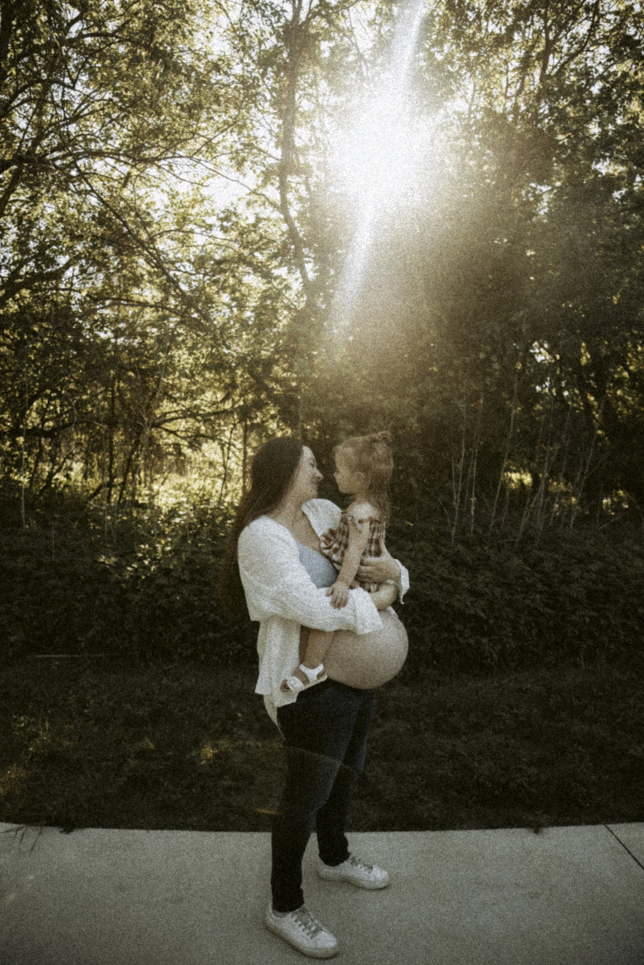 A woman is holding a young girl in her arms outdoors during sunset. The woman has long dark hair and is wearing a white dress, and the girl has light hair and is wearing a plaid shirt and white sandals. They are surrounded by trees with sunlight filtering through the leaves.