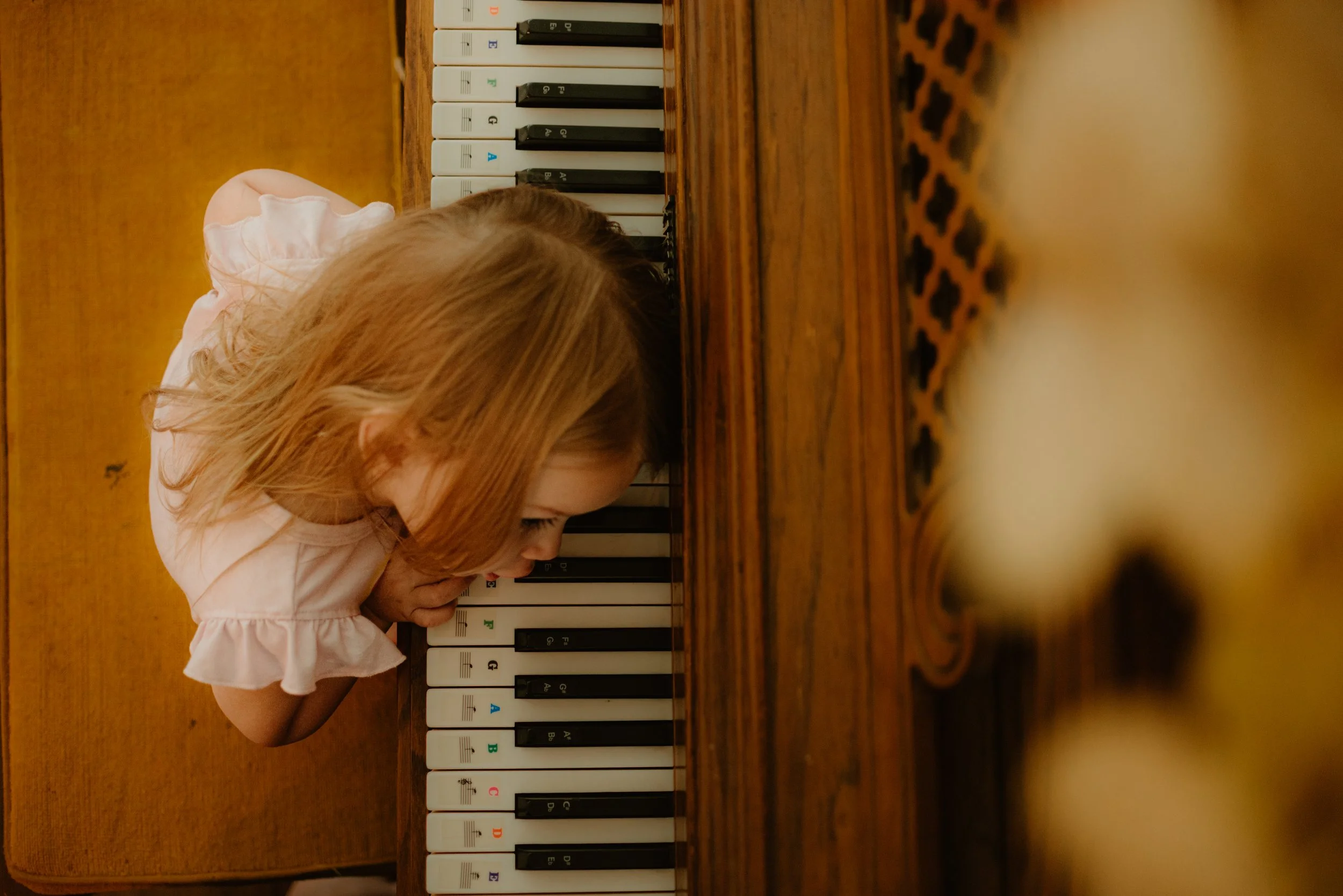 A young girl with red hair leaning over a wooden table, playing on a piano. The piano has black and white keys with some colored labels. The background features a yellow wall.