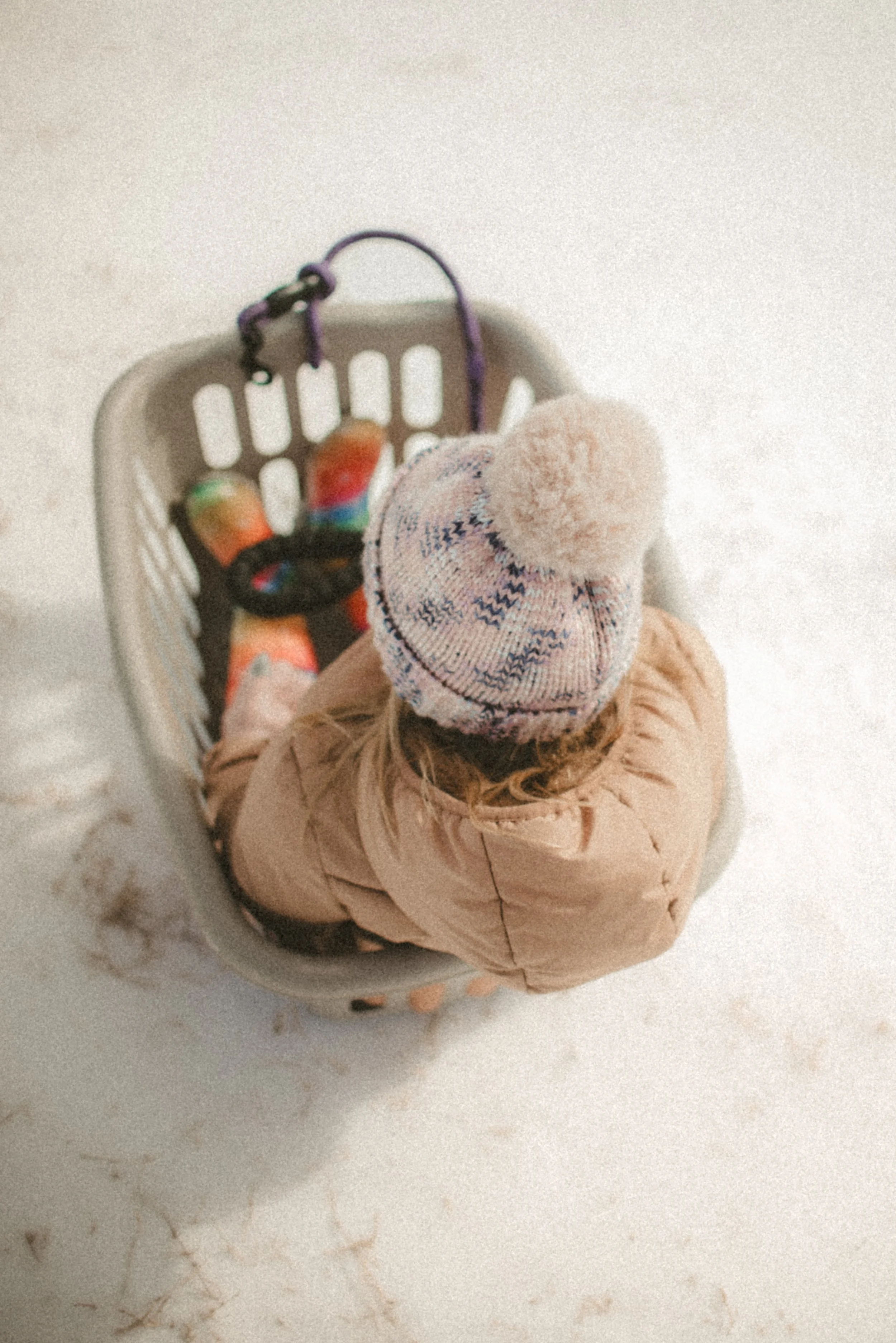 Child wearing a pink puffy coat and a colorful knit hat with a pom-pom, sitting inside a shopping cart with multicolored toys, on a white marble surface.