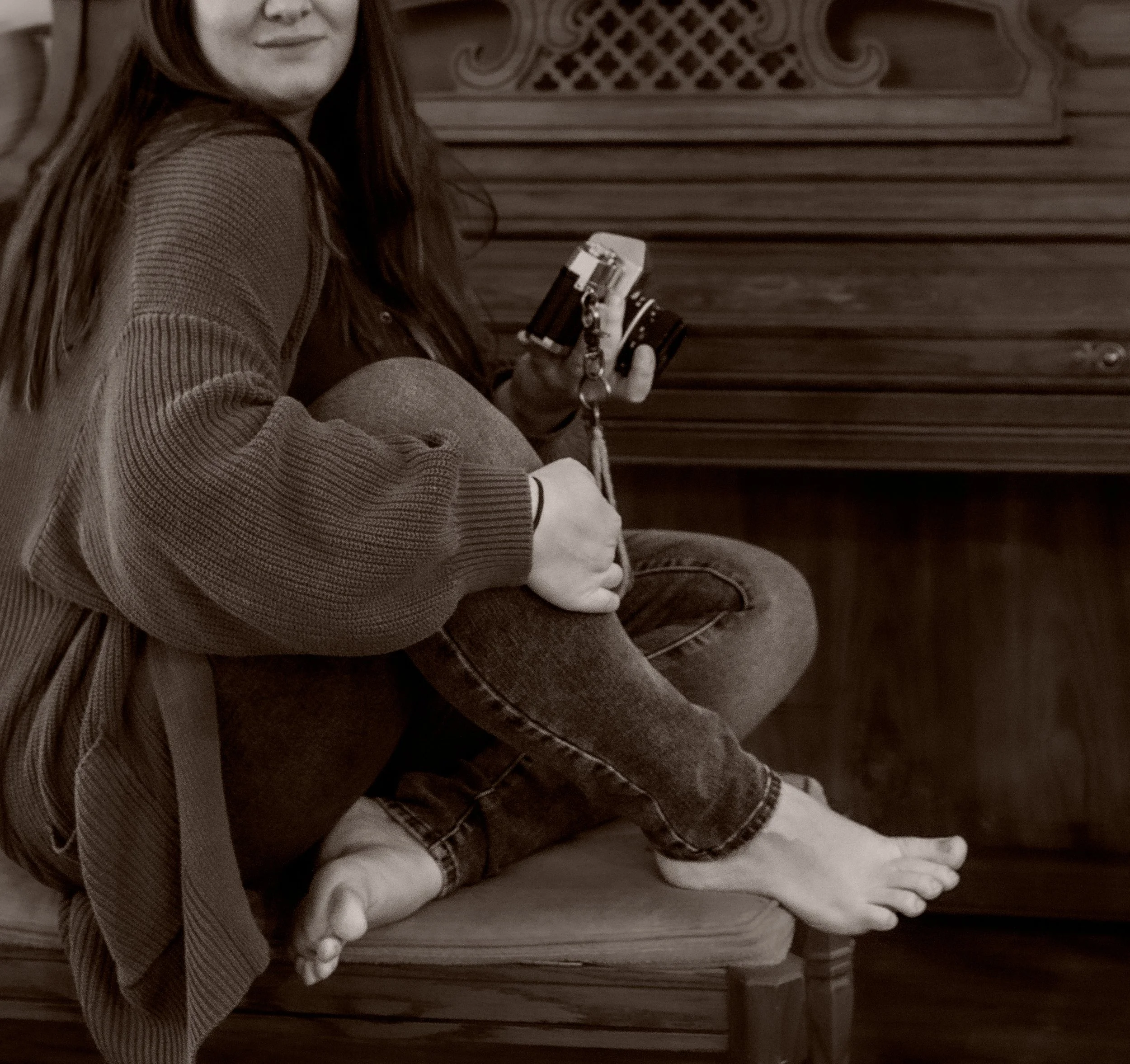 A person sitting on the ground holding a camera, with their legs crossed and wearing a striped long-sleeve shirt and jeans. The person is positioned in front of a carved wooden structure.