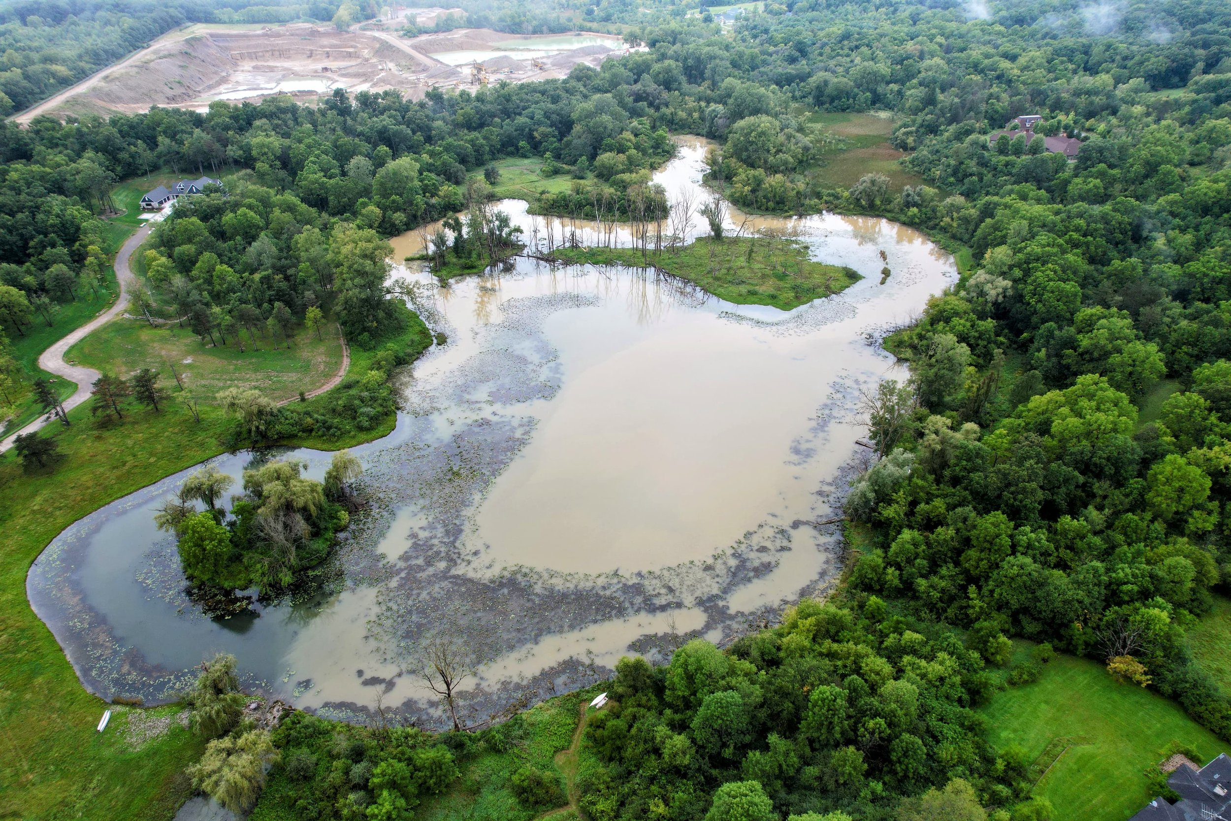 Aerial view of a lake with surrounding green trees and a grassy landscape, indicating a wetland or flooded area, with some houses and a construction site in the background.