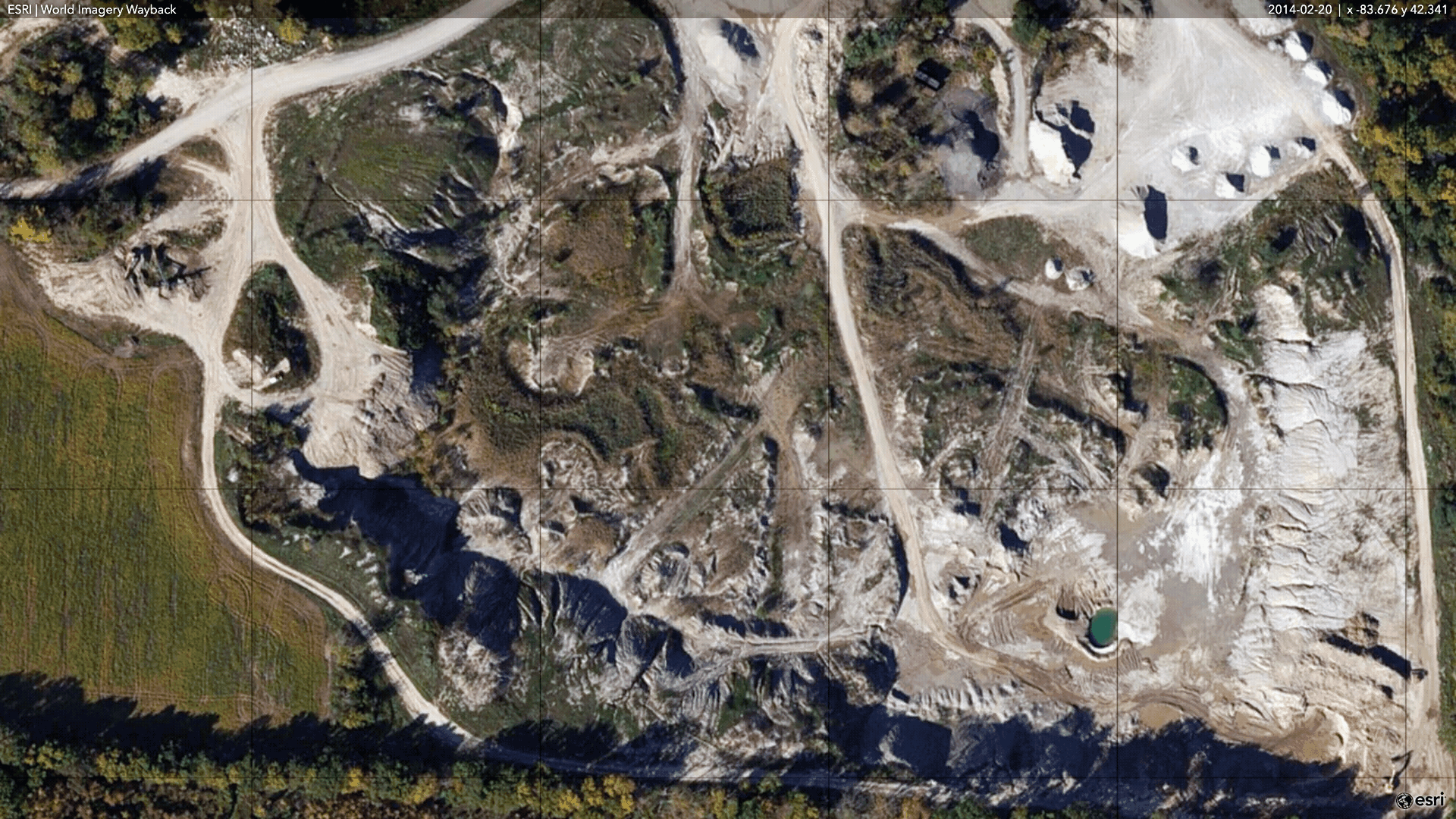 An aerial view of a quarry or open-pit mining site with exposed rocky and sandy terrain, scattered pools of water, and winding dirt roads, surrounded by forested areas.