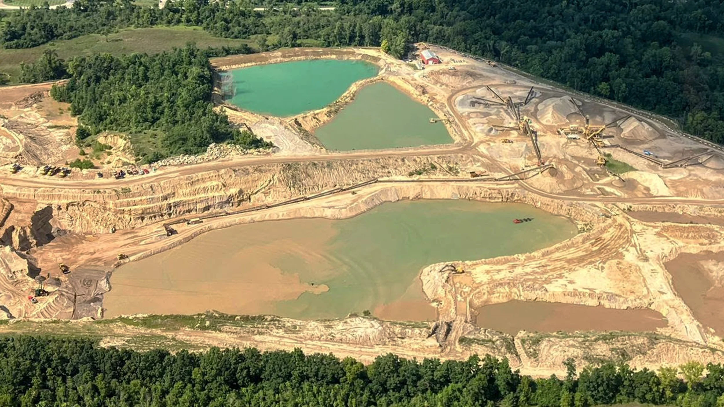 An aerial view of a mining operation in a forested area, showing multiple large open pits with water-filled sections, construction equipment, and dirt roads.