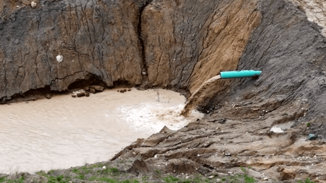 Water flowing from a pipe on a rocky cliff into a muddy area below.