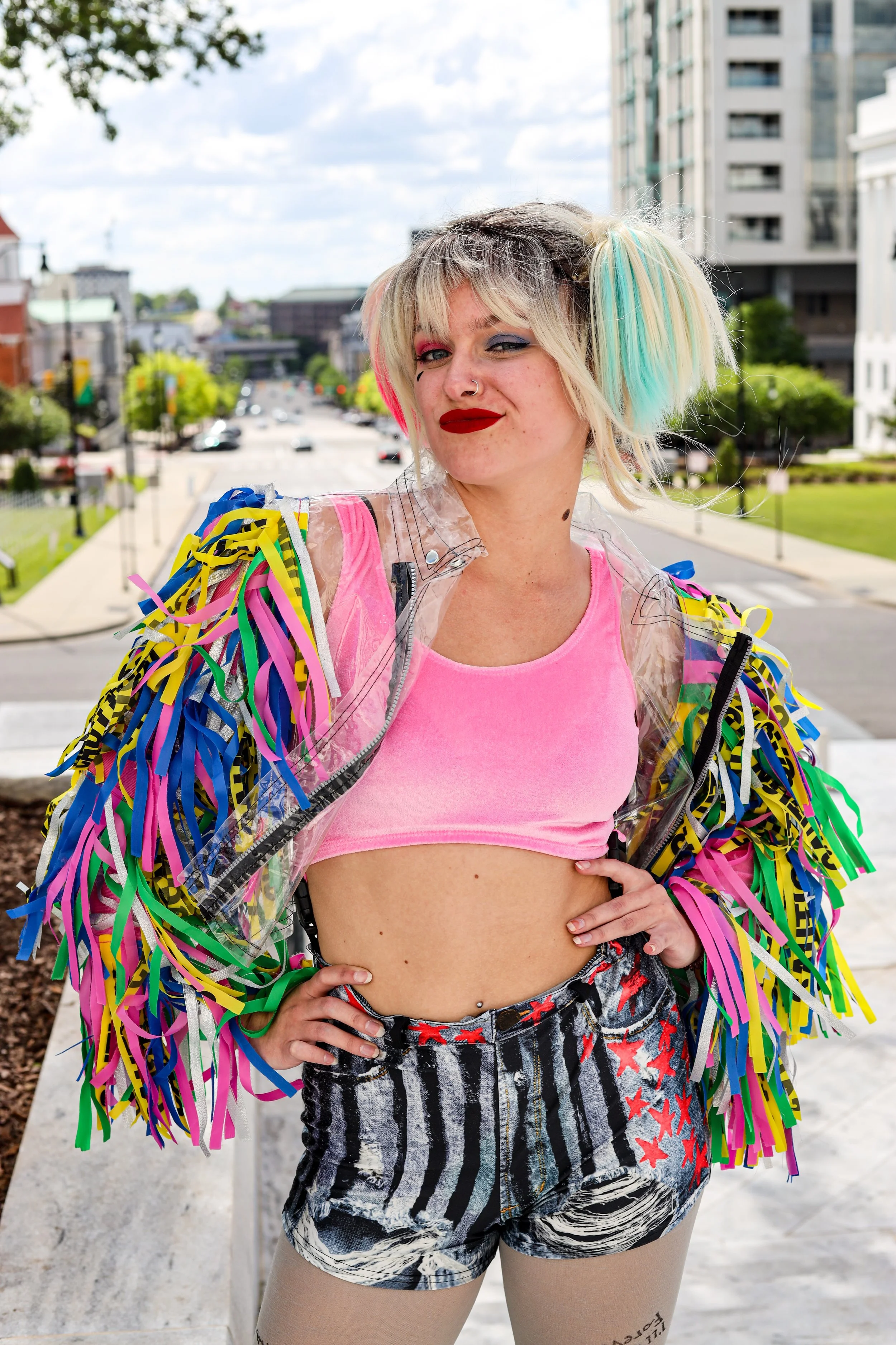 Person with blonde and turquoise hair, bold makeup, and colorful fringe jacket posing on a city street