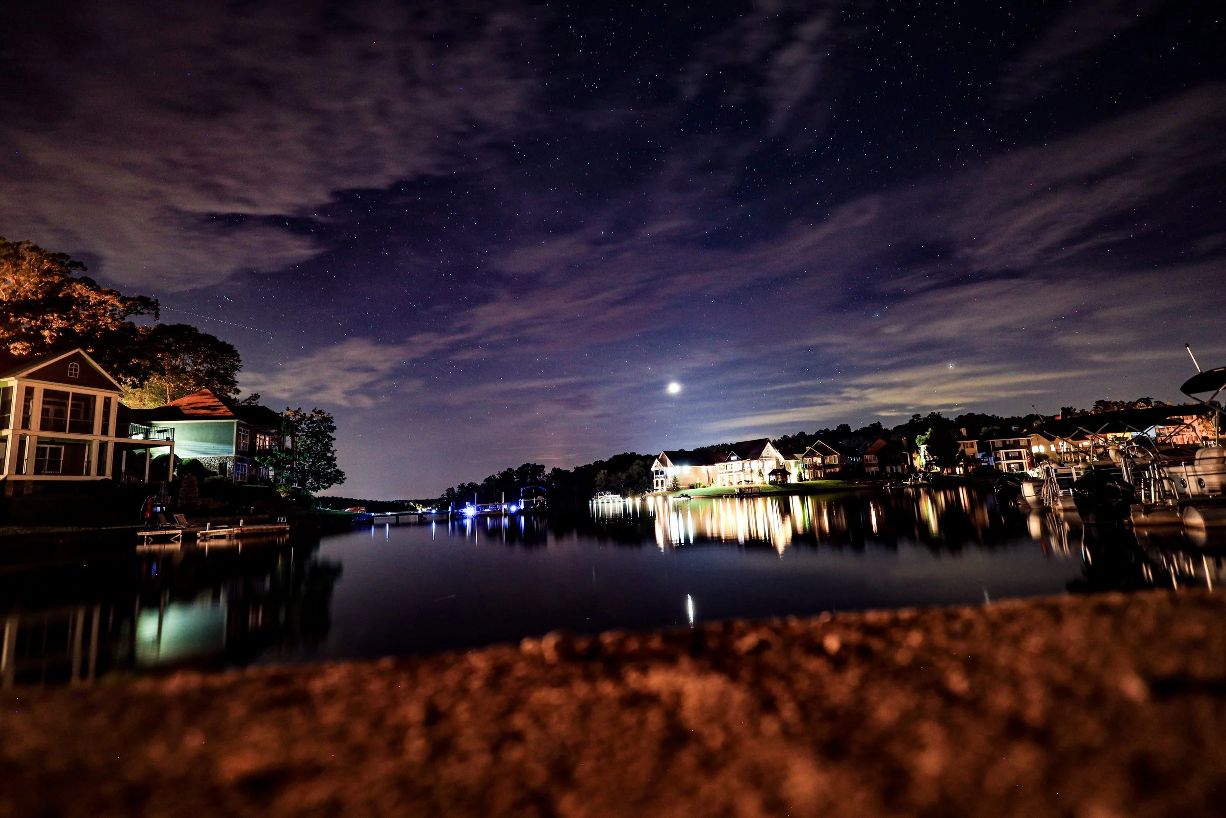Star-filled night sky over illuminated waterfront homes, docks, and boats reflected in calm water