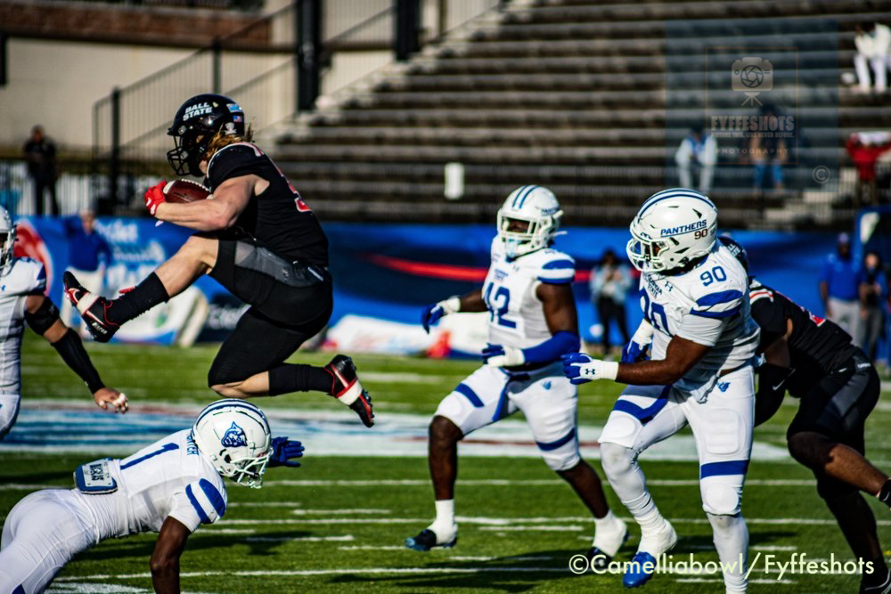 College football game with a black-uniformed player leaping over a diving defender as white-and-blue Panthers players pursue.