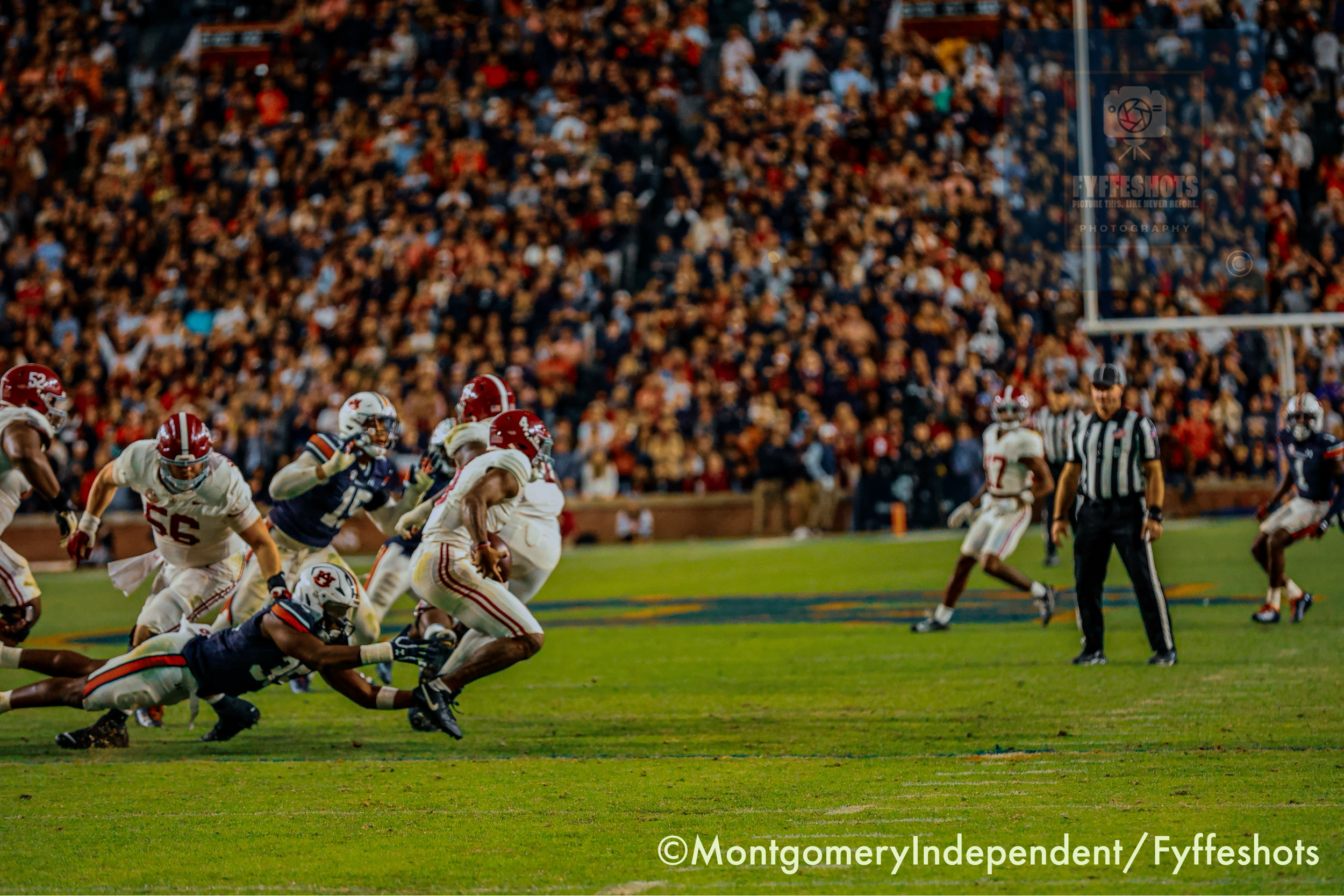 College football players compete on a field in front of a packed stadium crowd, with a referee watching the play.