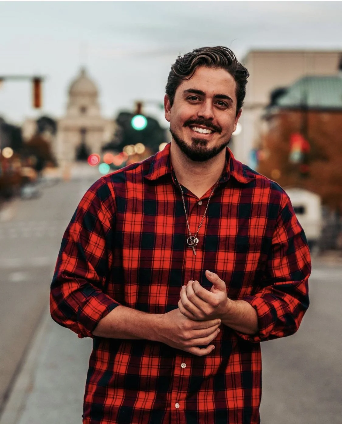 Smiling bearded man in a red plaid shirt standing on a city street with blurred traffic and buildings behind him