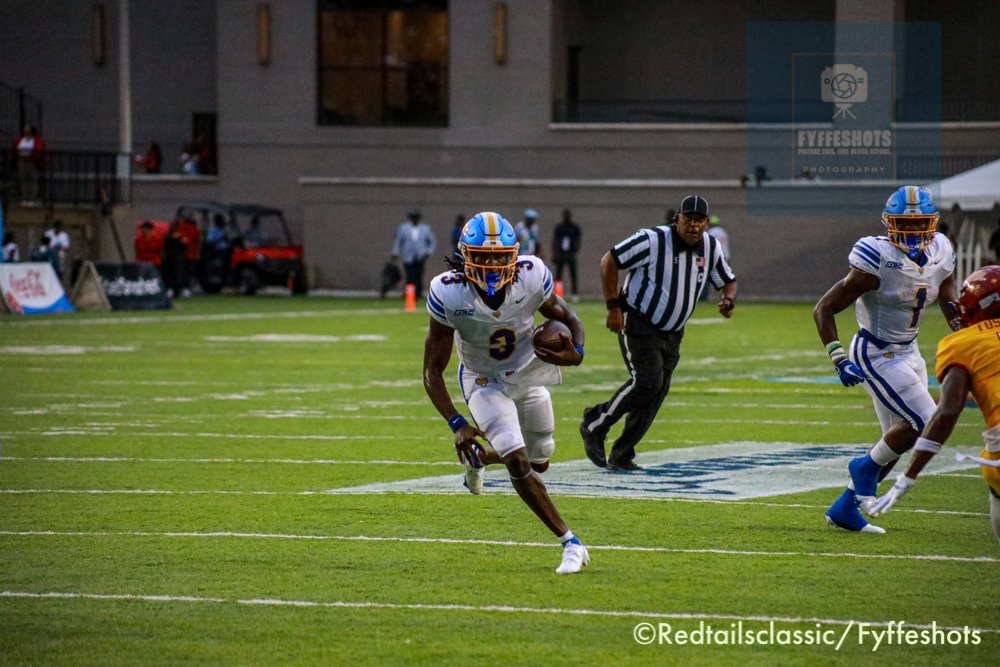 College football player in a white and blue uniform runs with the ball as teammates and a referee follow on the field.