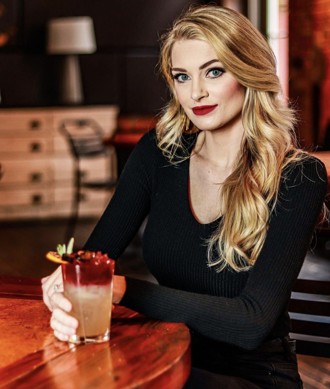 Blonde woman in a black top holding a fruit-garnished cocktail at a restaurant table