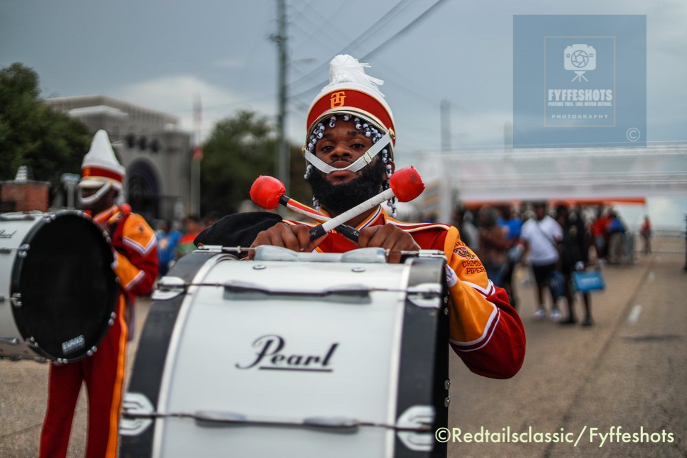 Drum major in an orange marching band uniform and white plumed hat playing a Pearl bass drum in a street parade