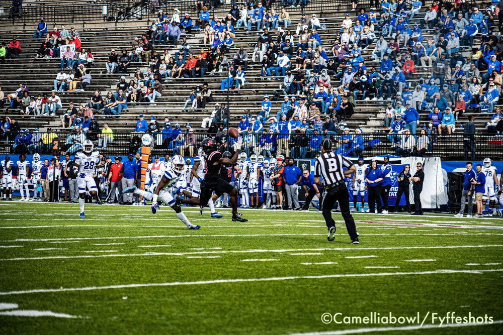 College football player in black catches a pass as defenders in white chase him on the field, with a referee and crowded stadium stands behind