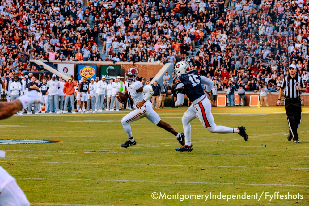 College football quarterback runs with the ball as a defender gives chase in front of a packed stadium crowd.