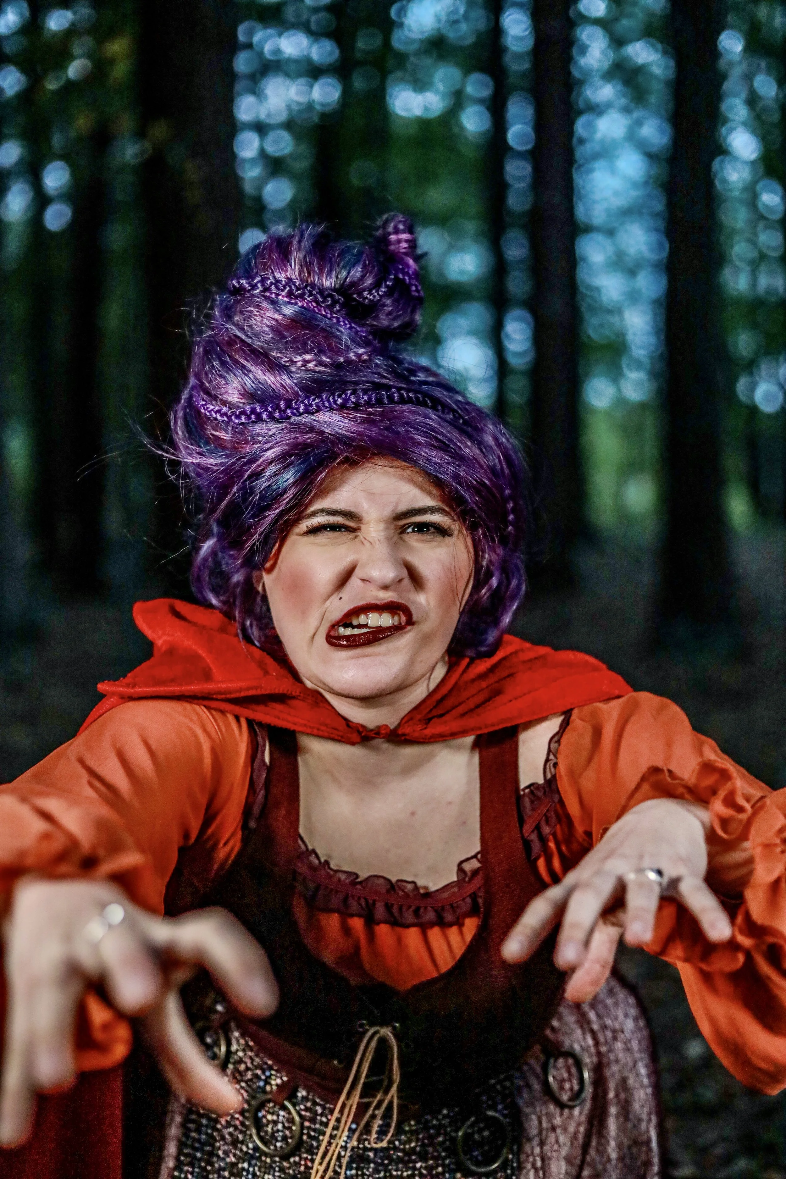 Smiling person with purple hair and braided updo wearing a red and orange costume against a dark, blurry forest background