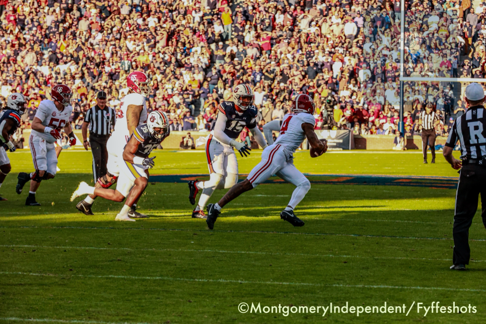 College football game with a white-uniformed ball carrier sprinting past defenders on a sunlit field before a packed stadium crowd