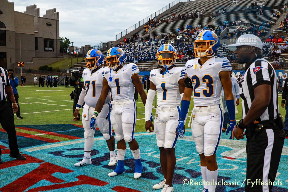 Four college football players in white and blue uniforms stand on the field with a referee, band, and spectators in the stadium behind them.