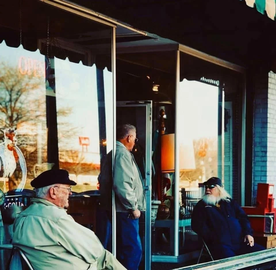 Three older men outside a storefront café, with two seated beneath awnings and one standing in the doorway.