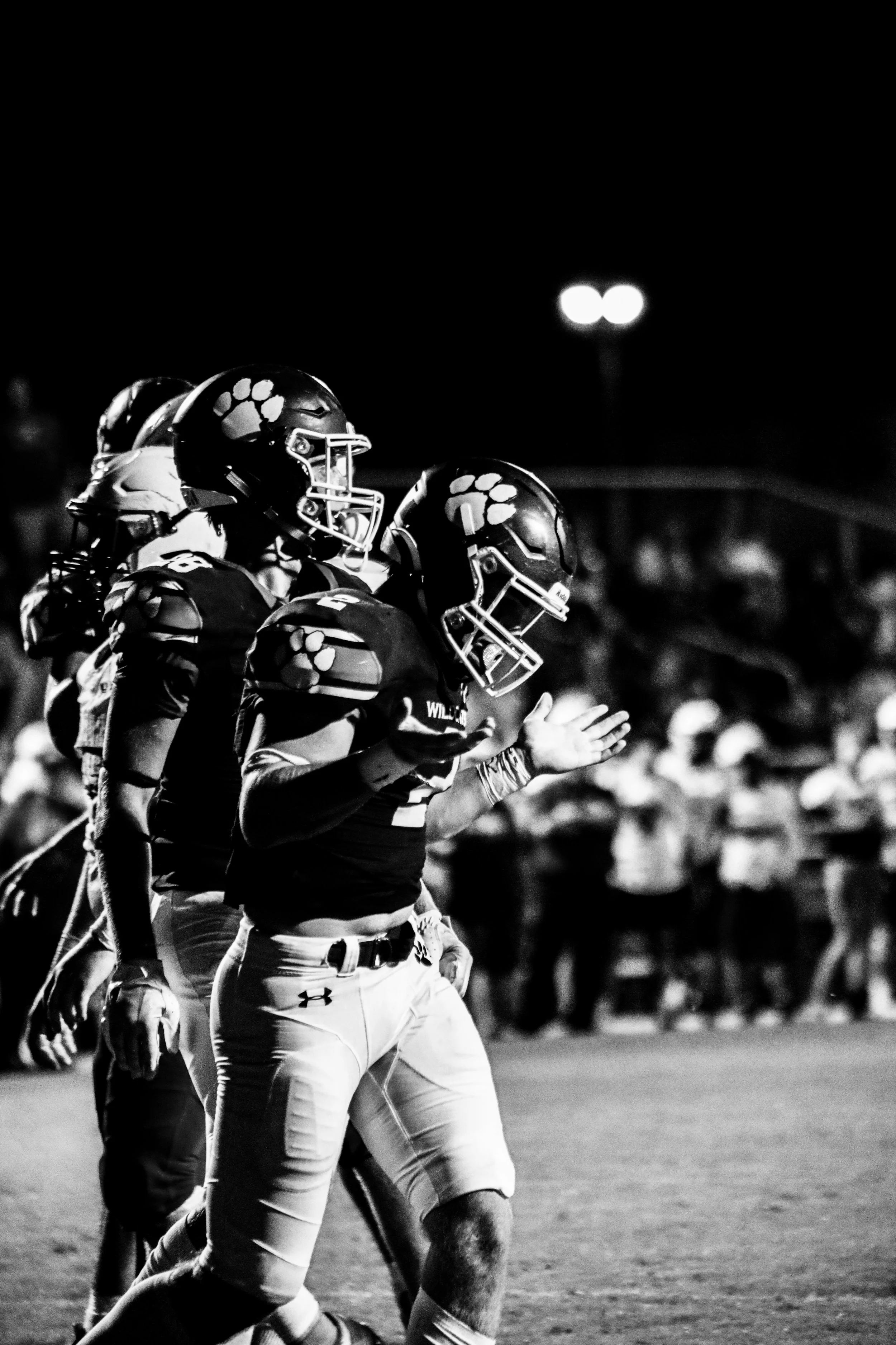 Black-and-white photo of American football players in helmets and uniforms celebrating on a nighttime field.