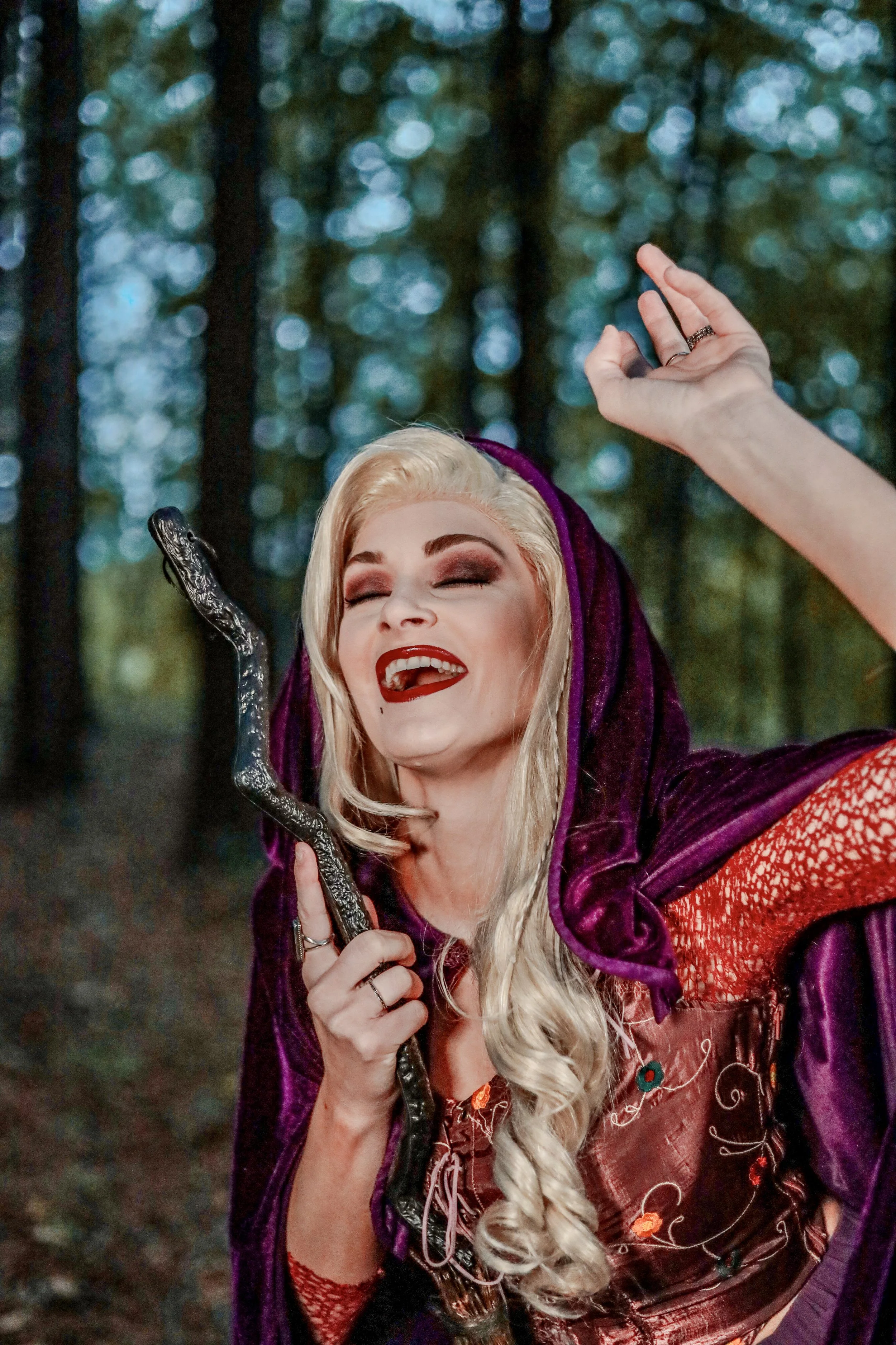 Smiling blonde woman in a purple hooded costume holding an ornate black staff in a misty forest