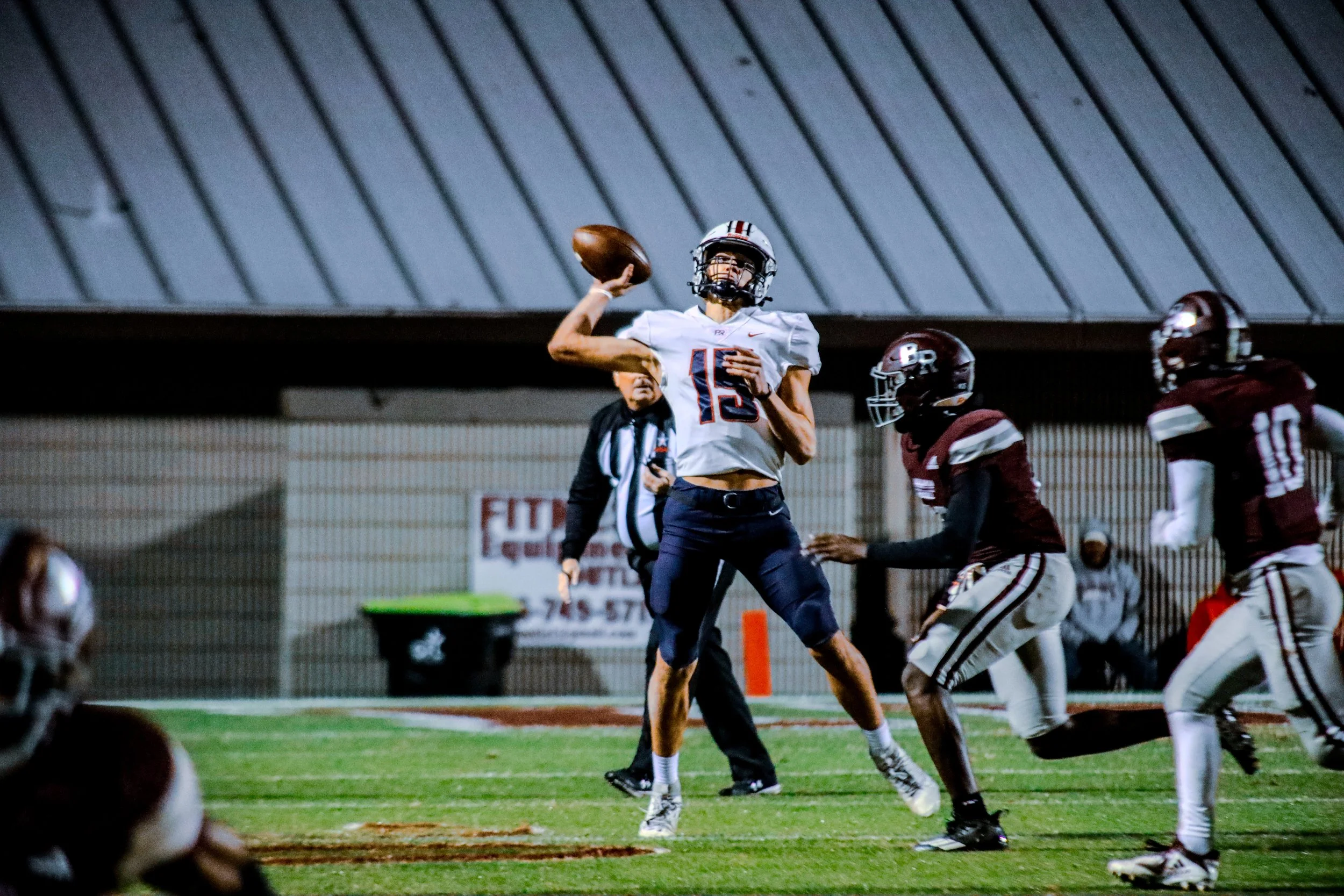 High school football quarterback in a white No. 15 jersey throws the ball as defenders pursue him on the field.